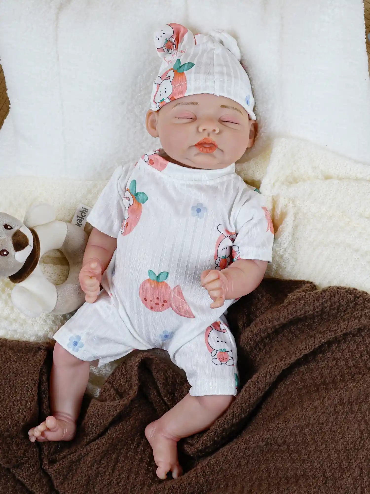 Lifelike reborn baby doll dressed in a white, fruit-print romper and a matching "bunny" hat, lying next to a plush koala toy, with a teething ring nearby
