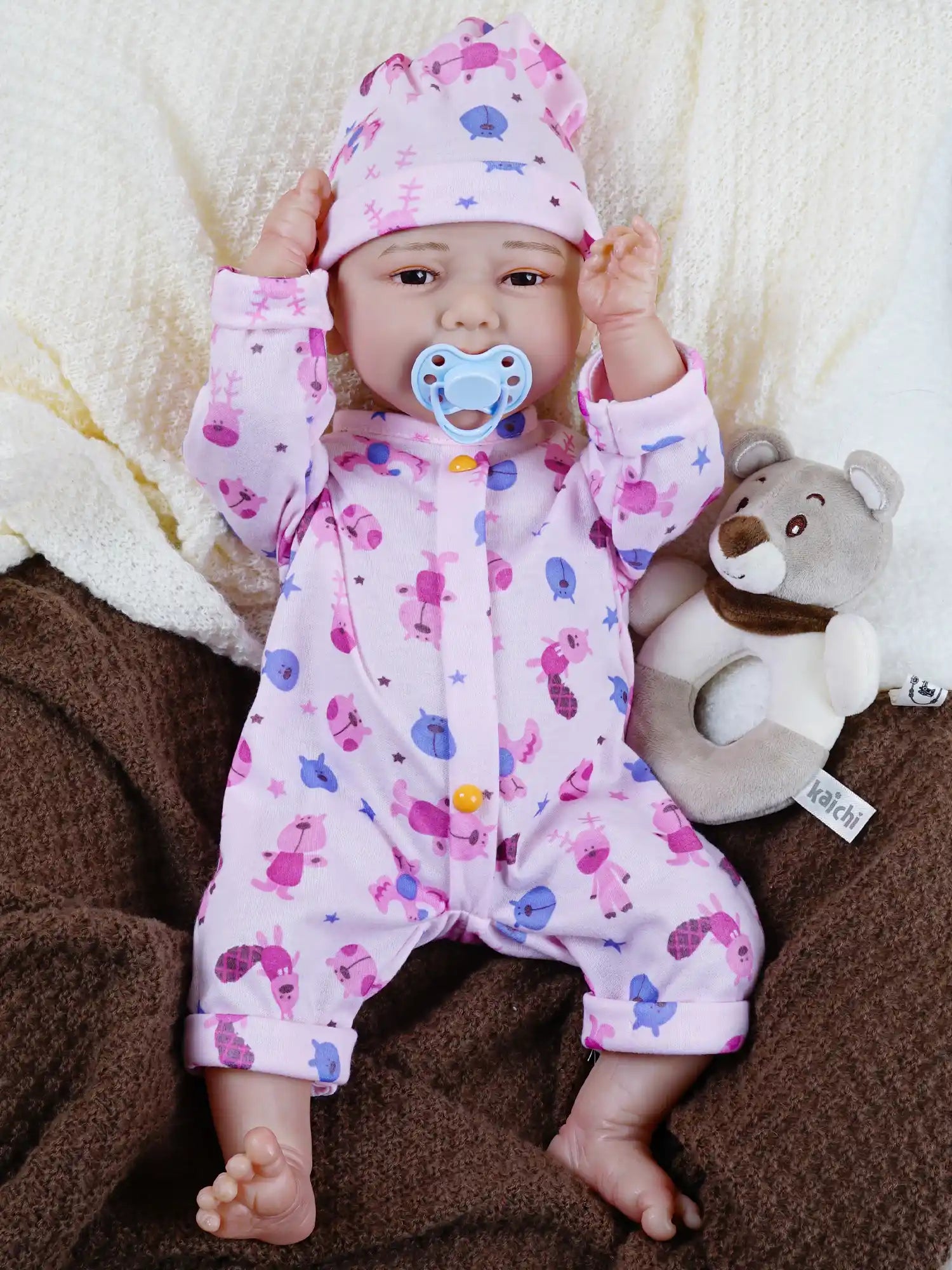 Smiling reborn baby doll in pink sea-themed pajamas seated with plush bear toys, on a cream blanket beside a brown blanket