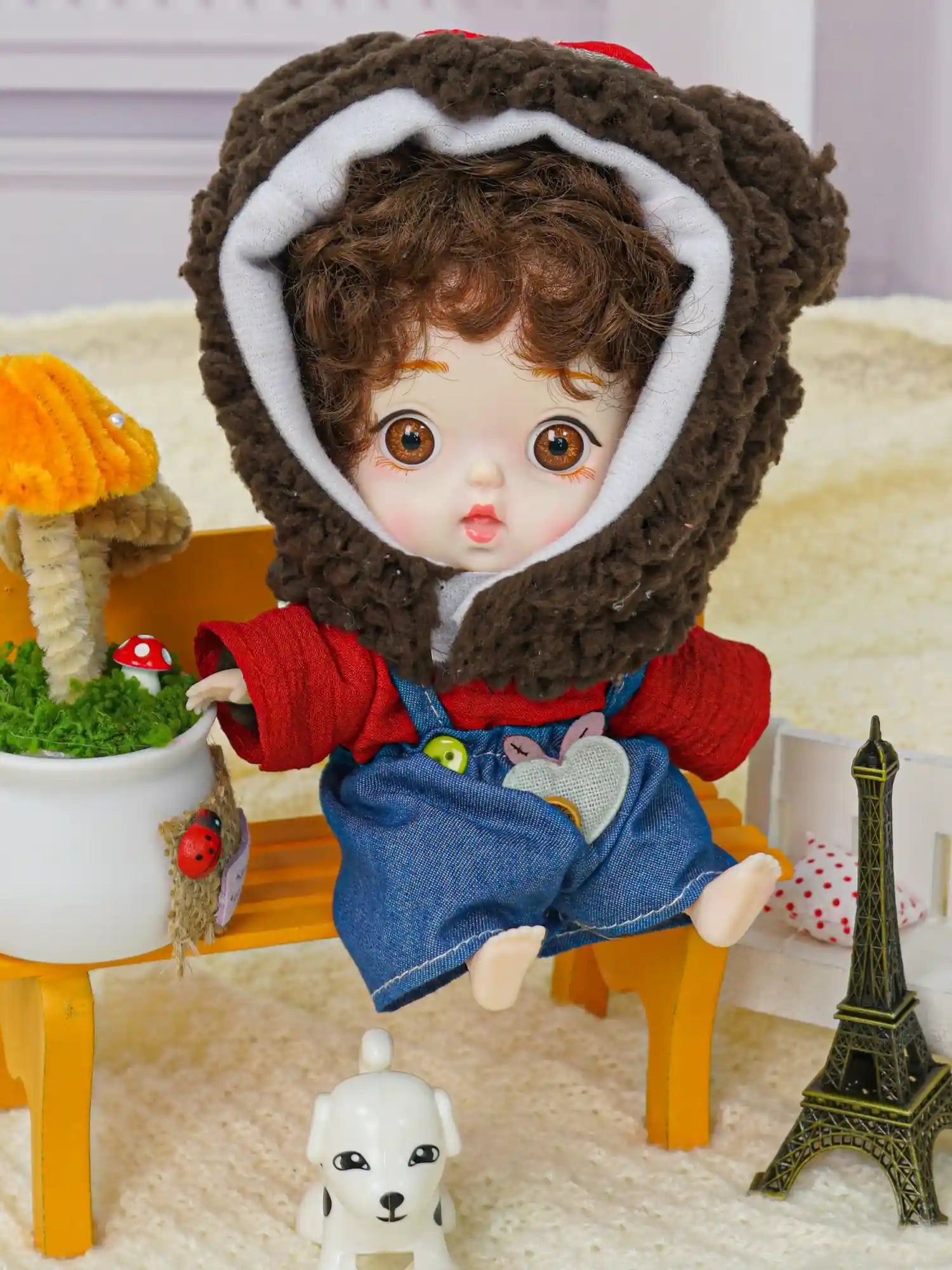 A ball-jointed doll sporting a hood with bear ears, sharing the stage with a miniature Eiffel Tower and a white puppy figure.