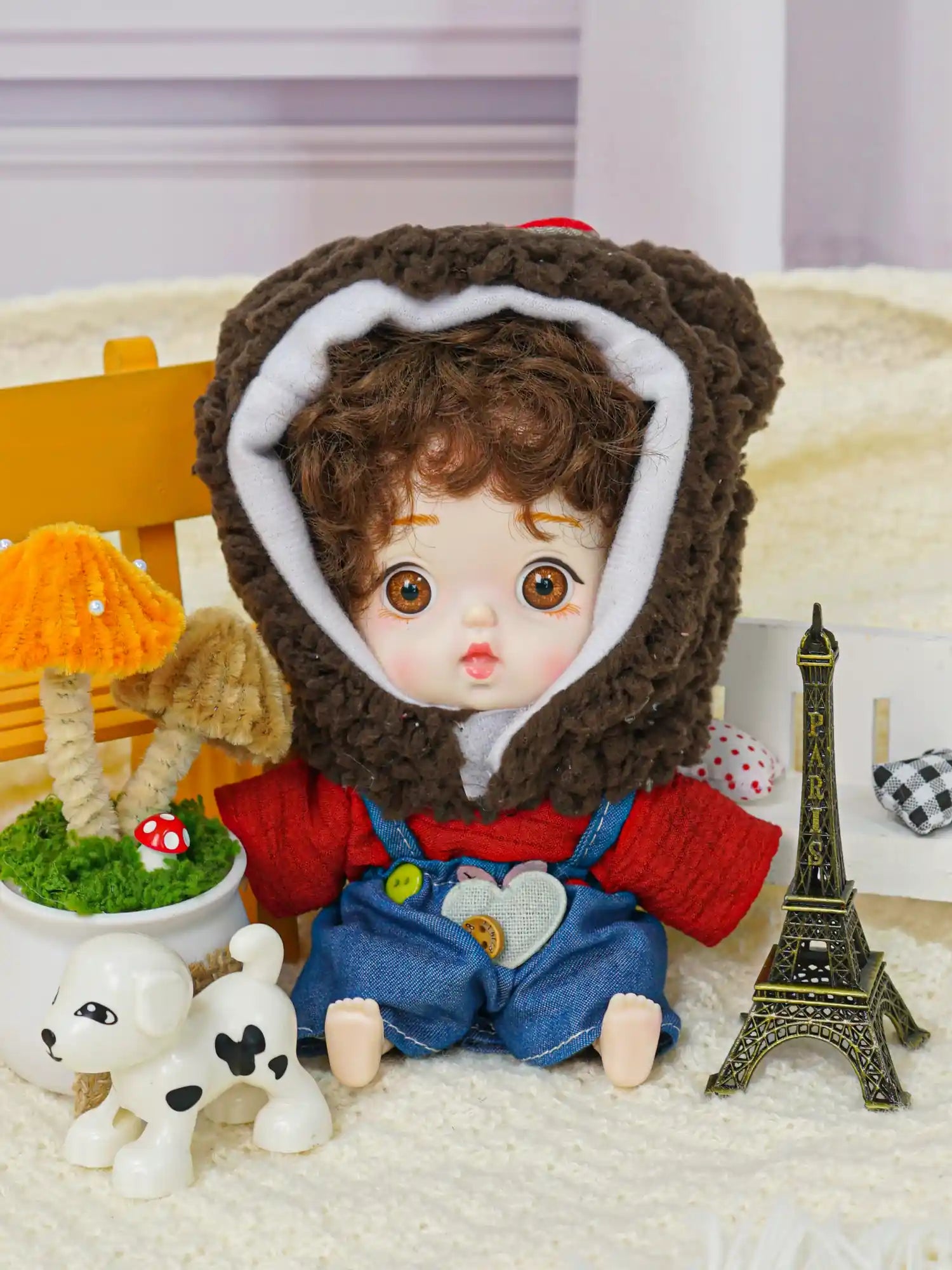 A brown-eyed doll with a plush bear hood and overalls, next to a small Eiffel Tower and a playful dog figurine.