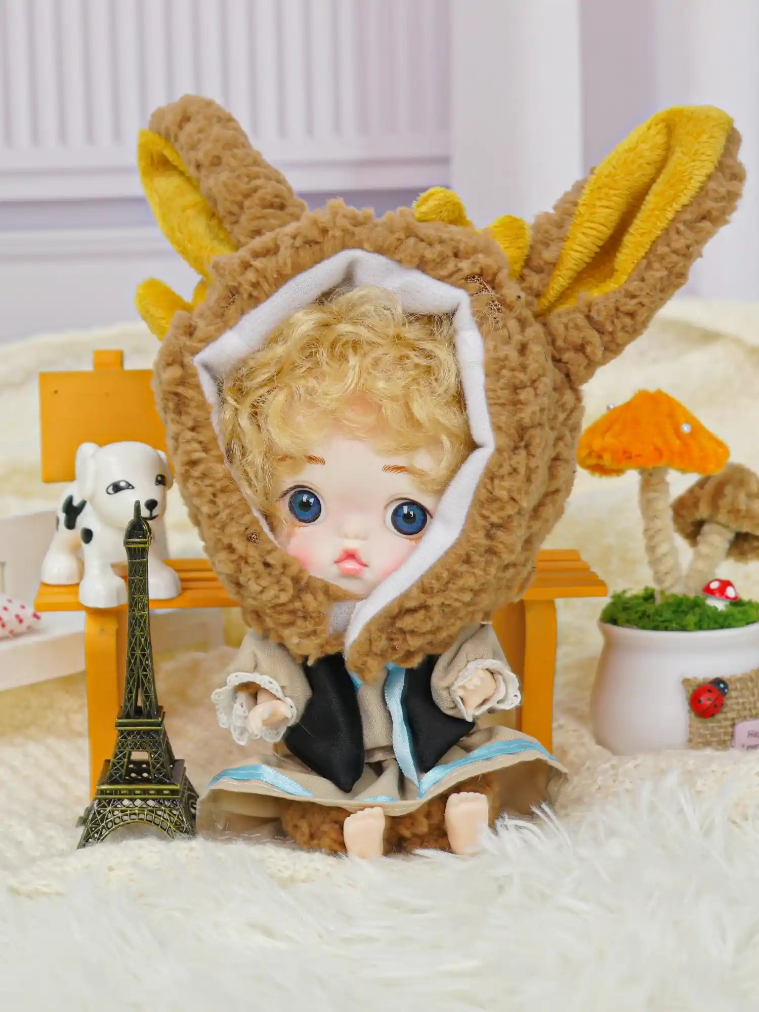 An adorable doll with blue eyes and a cozy bunny hat, situated among playful miniatures including a tiny Eiffel Tower.