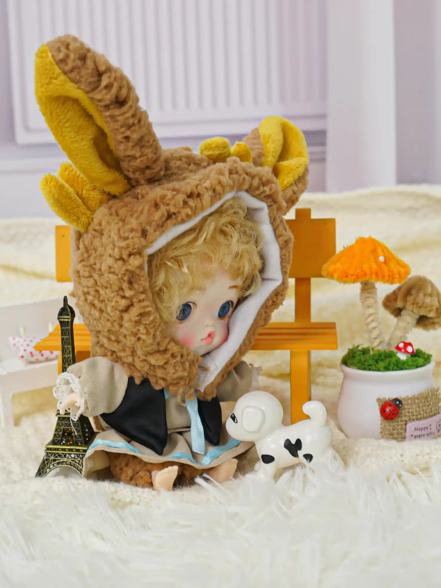 A doll with a plush rabbit headwear, nestled on a soft rug with a mini Eiffel Tower and a white dog companion.