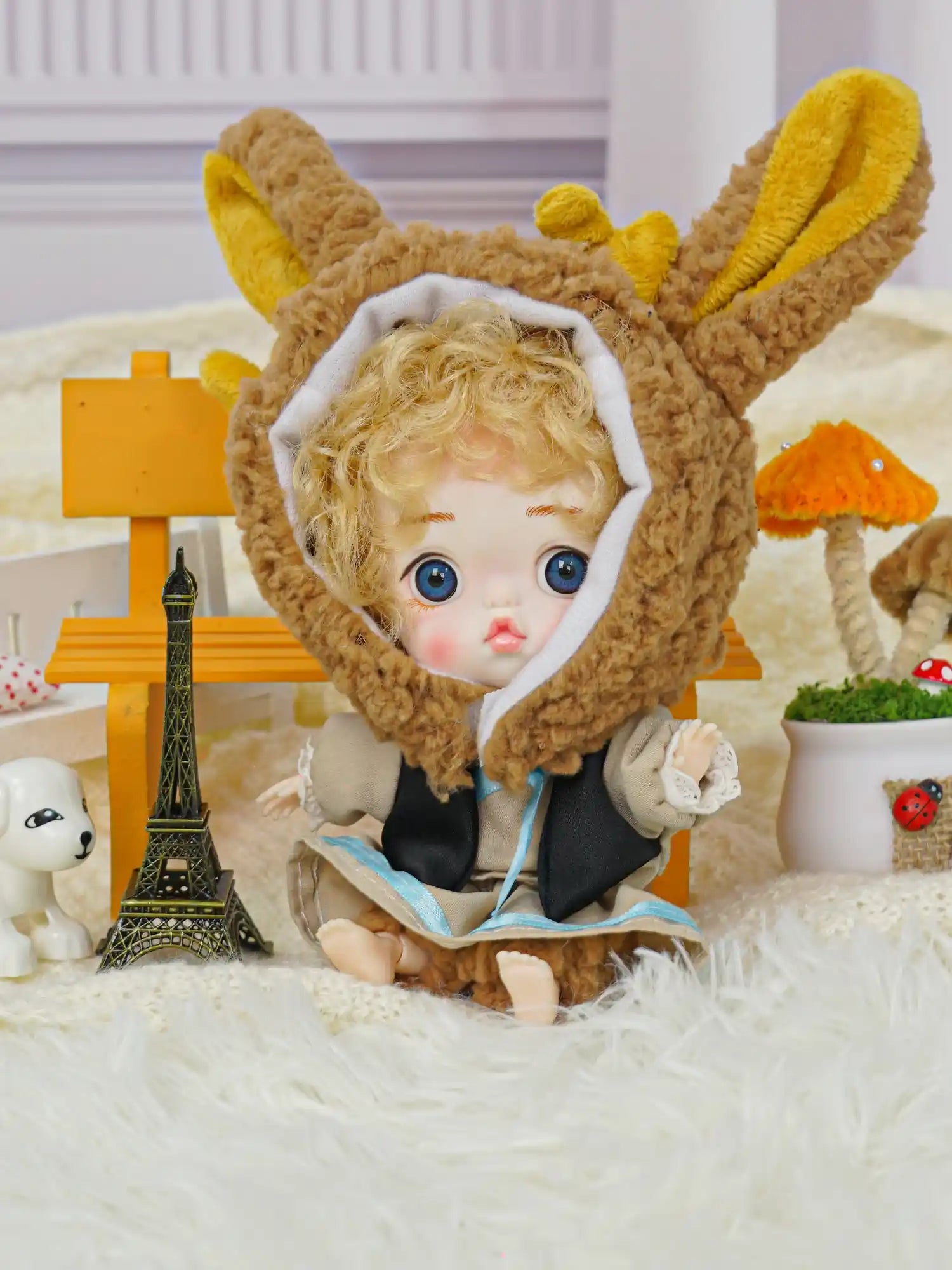 A whimsical ball-jointed doll with a rabbit hood and golden curls, surrounded by a toy Eiffel Tower and a small dog figurine.