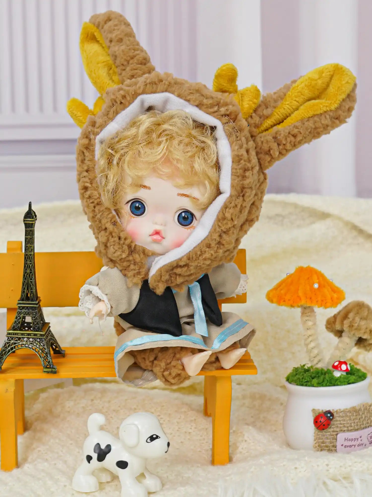 A blue-eyed doll with rabbit ears, posed with a small Eiffel Tower model and a toy canine on a plush background.