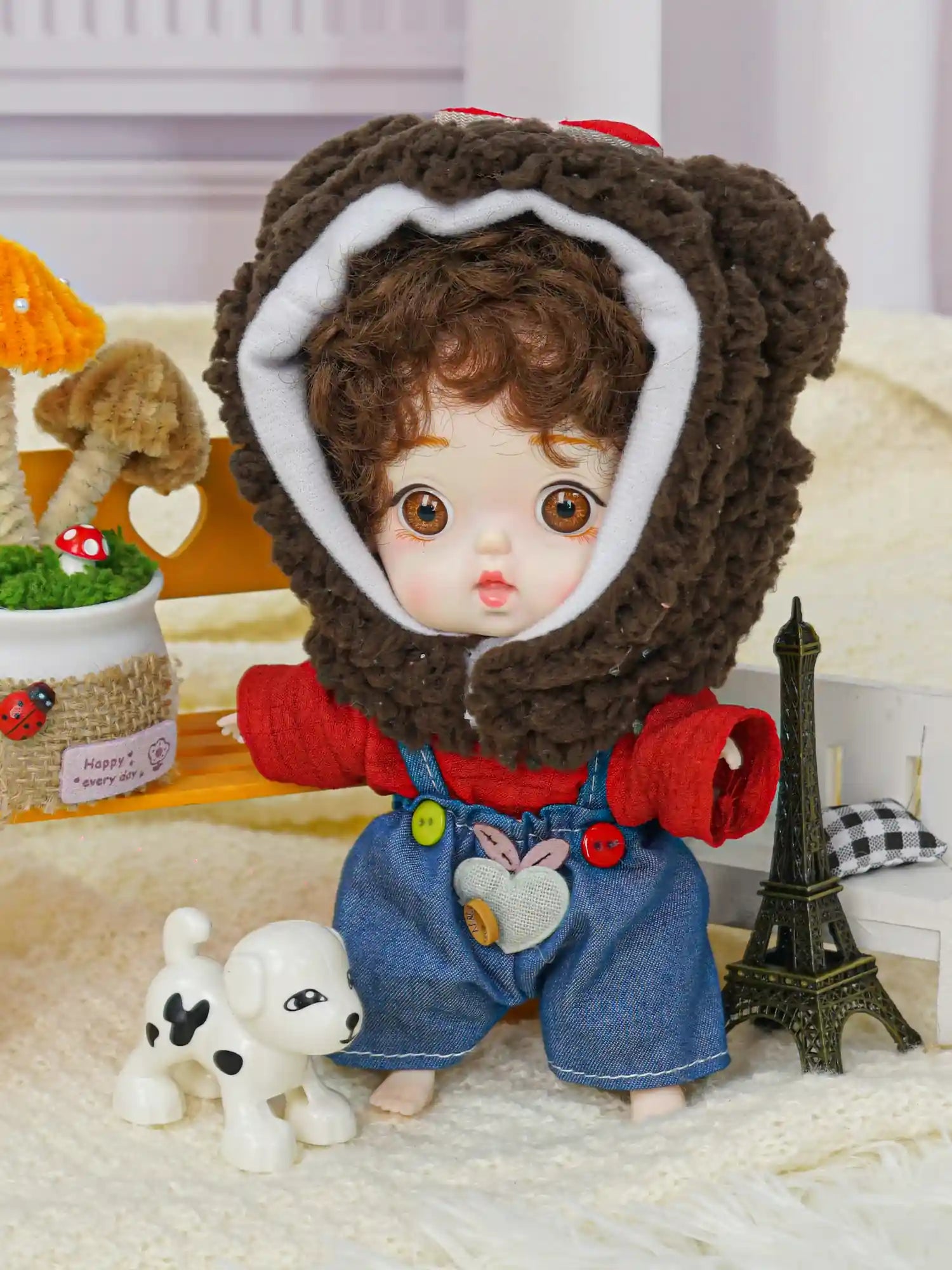 A ball-jointed doll with a teddy bear hat and bright eyes, posing with a mini Eiffel Tower and a small dog statue.