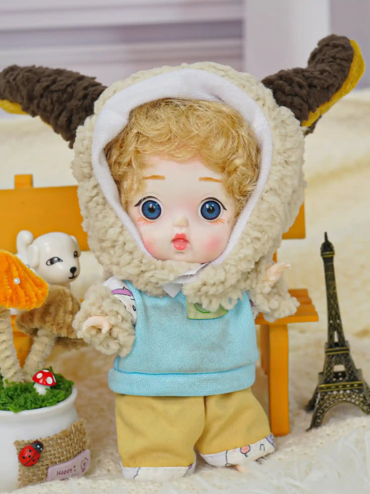 The BJD's captivating blue eyes shine from beneath a lamb's hood, with a miniature world around it.