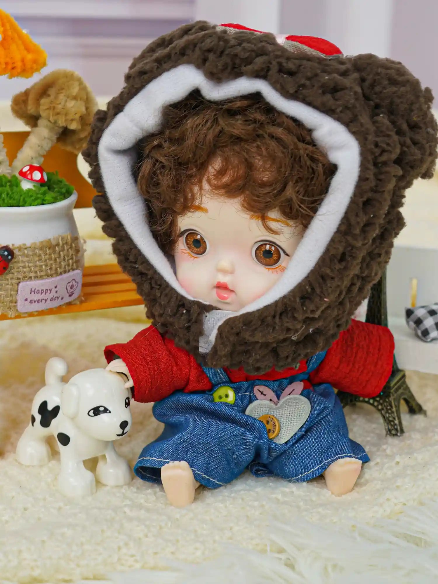 A cute doll with curly hair and a bear hood, standing next to a miniature Eiffel Tower and a white puppy toy.