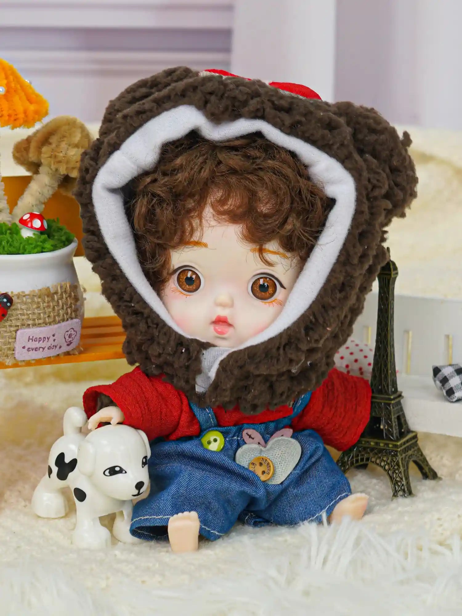 A brown-eyed doll wearing a cozy bear hood, displayed among a set of miniatures with a Parisian theme.