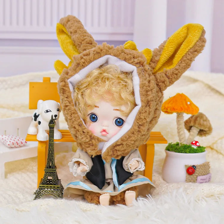 A whimsical ball-jointed doll with a rabbit hood and golden curls, surrounded by a toy Eiffel Tower and a small dog figurine.