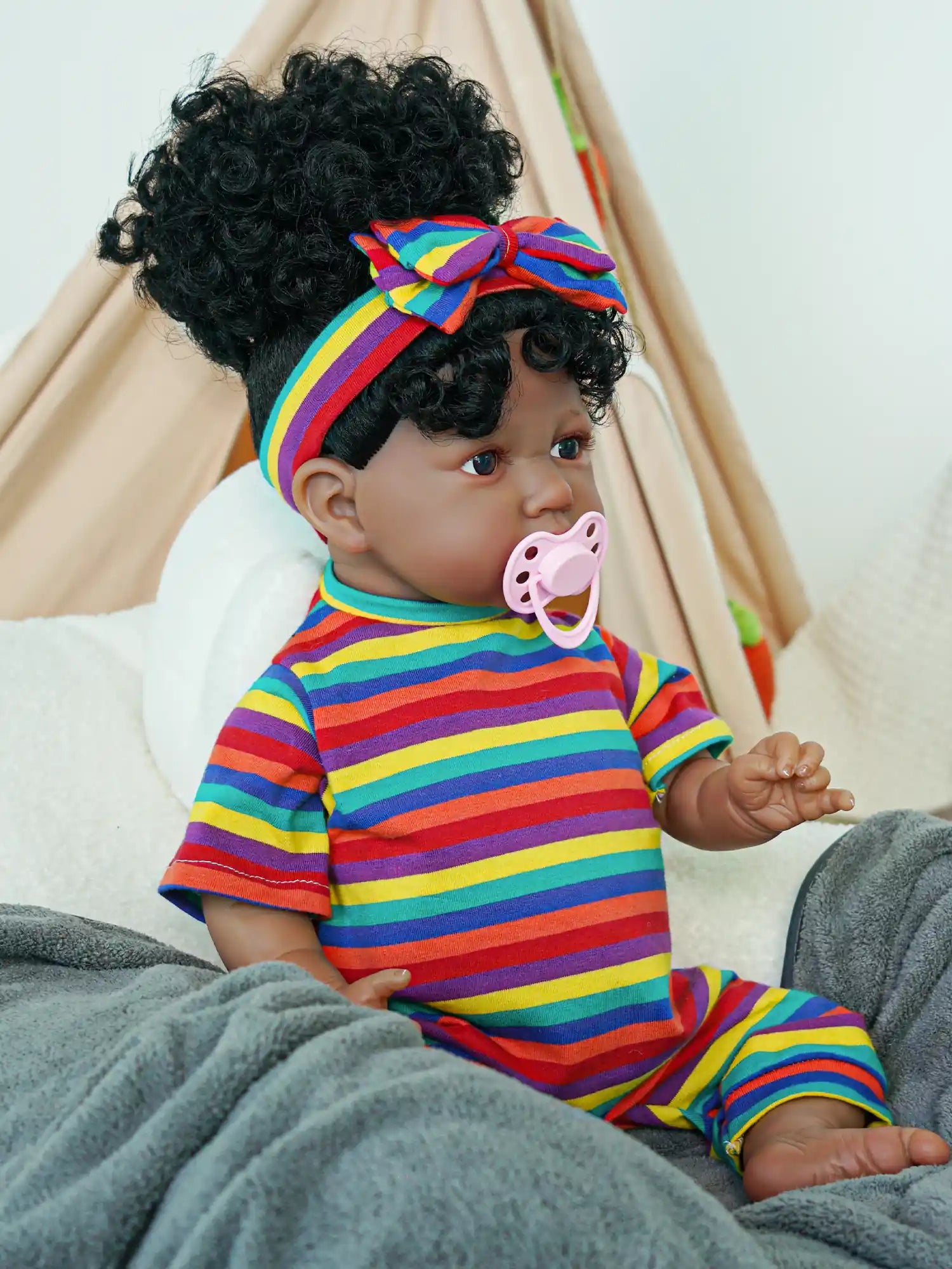 A black reborn baby doll sitting on a sofa wearing multi-colored striped clothes and having a magnetic pacifier in its mouth