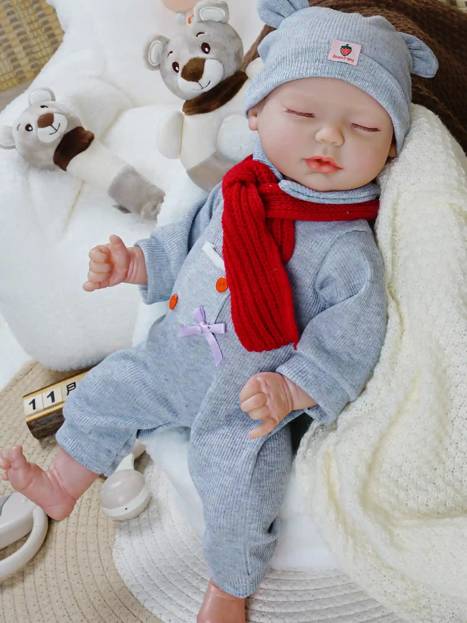 A baby doll dressed in a grey outfit with a matching hat and a red scarf, lying on a soft blanket. The doll's eyes are closed, and it is surrounded by plush bear toys, creating a cozy and peaceful scene.