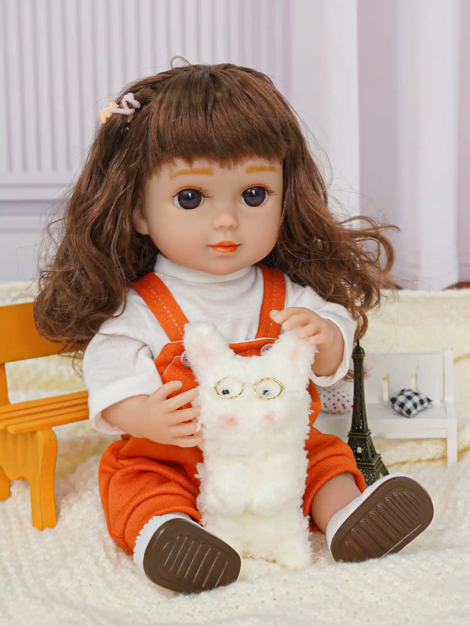 Doll with curly brown hair in orange overalls, white shirt, with plush dog and Eiffel Tower.