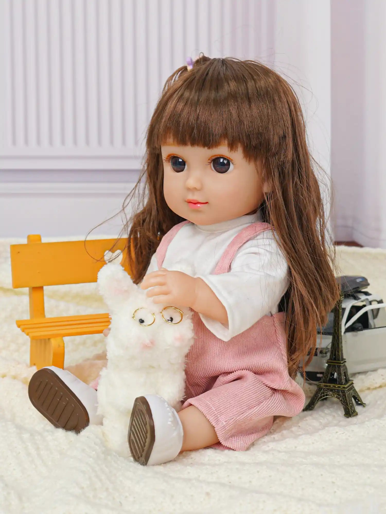 Doll with brown hair and pink overalls, waving, with a toy dog and Eiffel Tower.