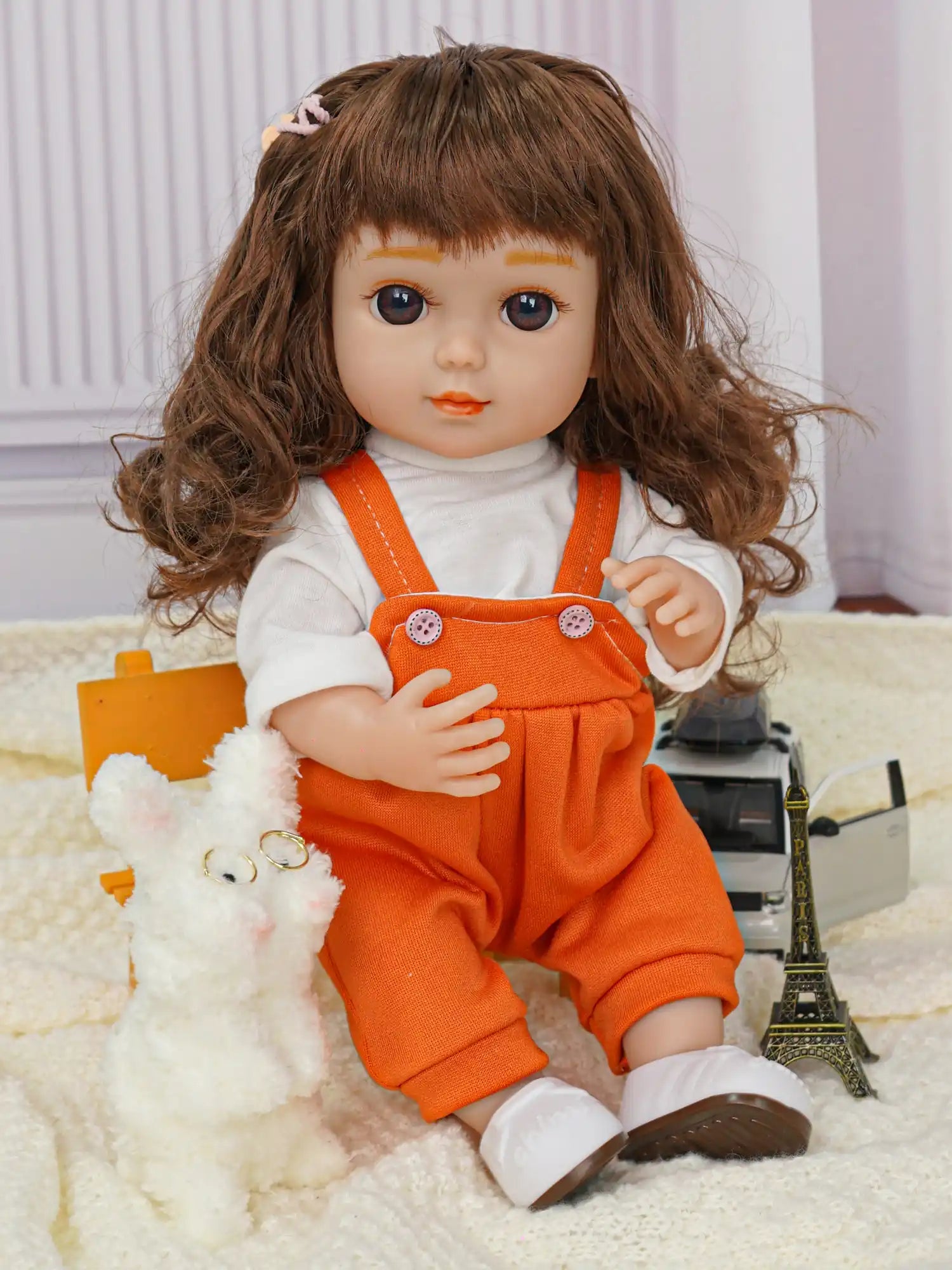 Curly-haired doll sitting, in a white top and orange pants, with a toy dog and miniature tower.