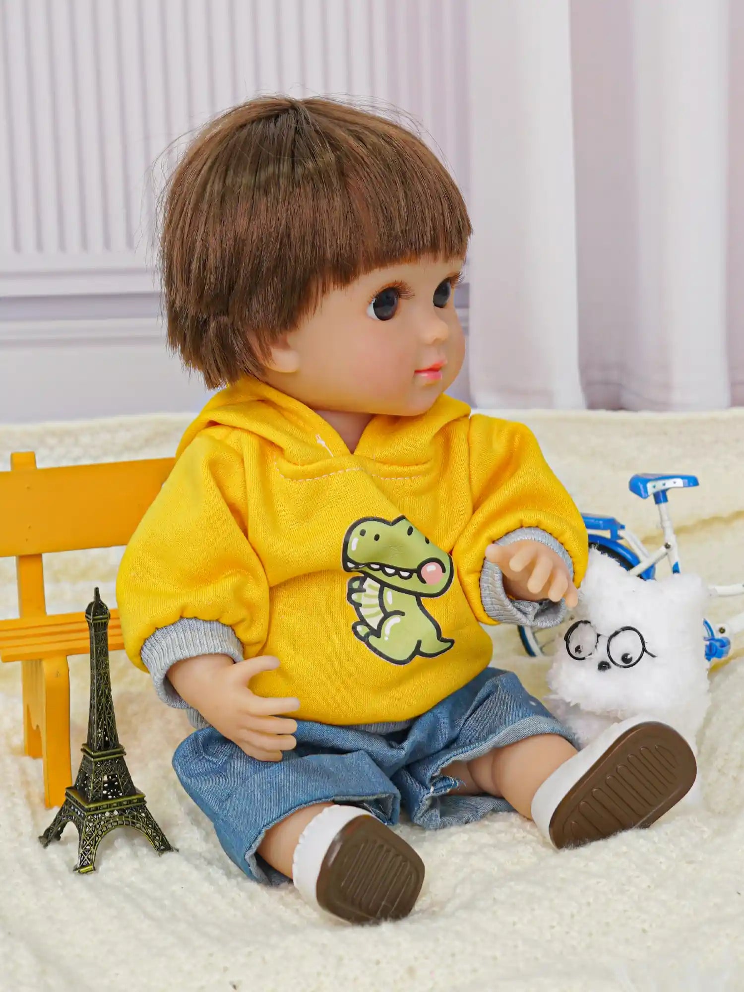 Childlike doll sitting with a yellow top, blue shorts, white dog toy, and a small bike.