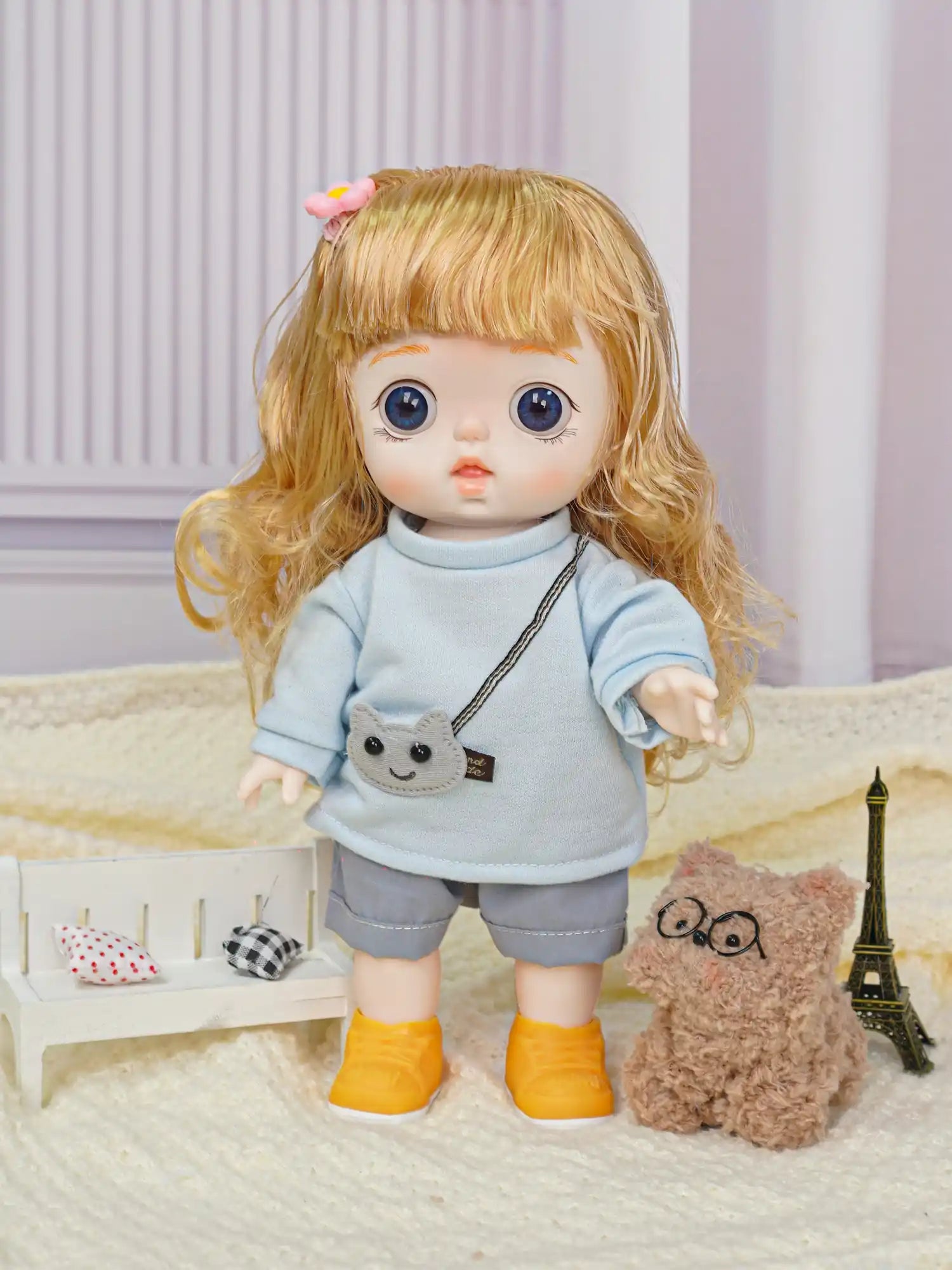 Doll with blonde curly hair and big blue eyes, dressed in a blue sweatshirt and grey shorts, accompanied by a plush toy.