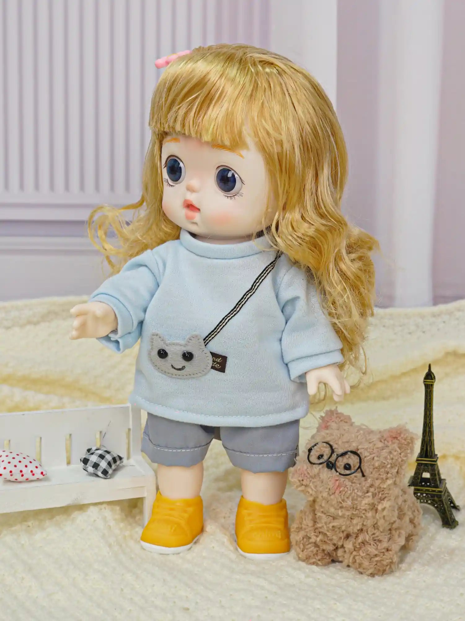 A little doll with wavy hair and a playful expression, wearing casual play clothes and standing near a soft, bespectacled toy.
