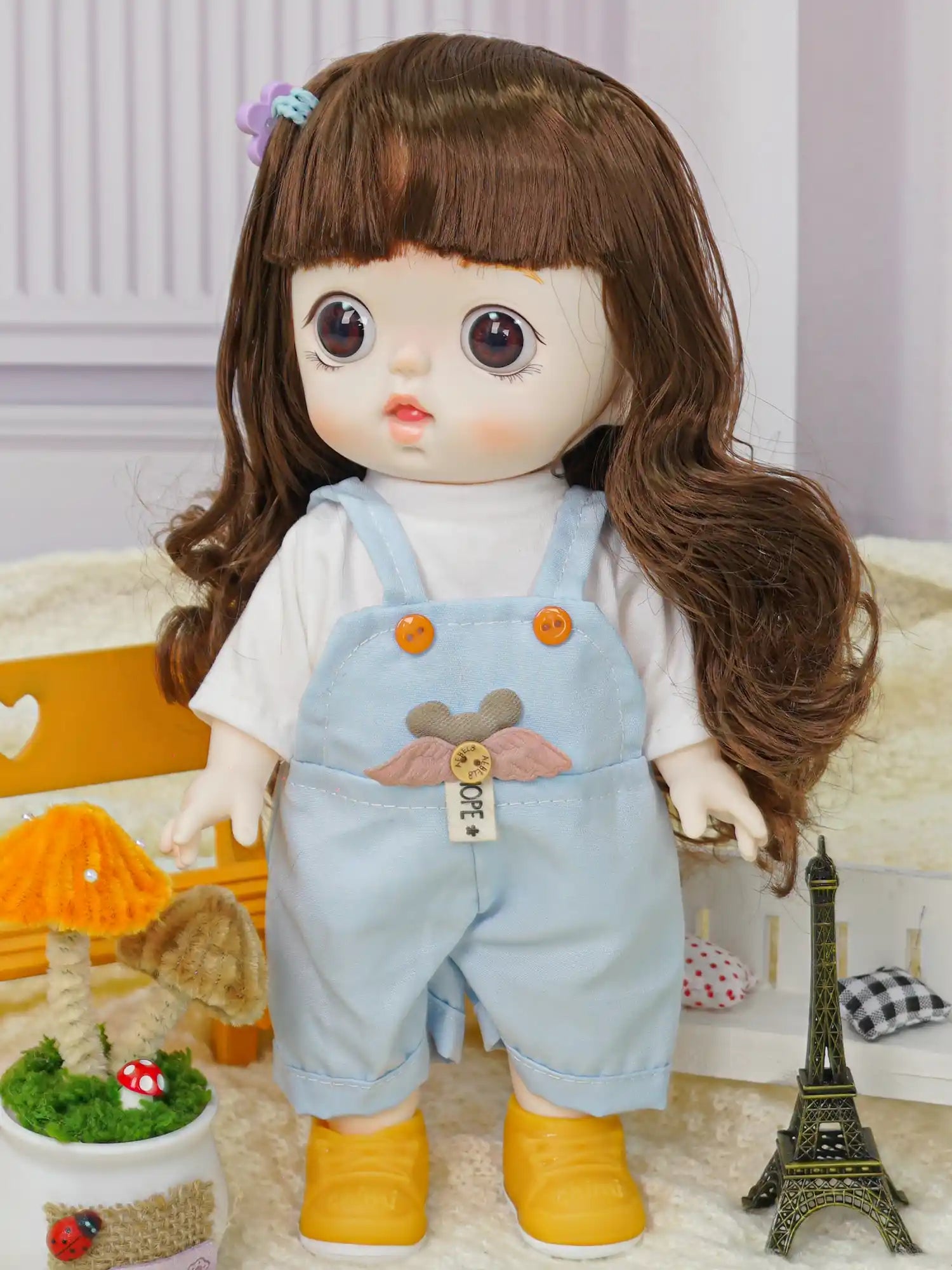 Doll with deep brown eyes and chestnut hair in a blue dungaree with a bear design, beside a miniature Eiffel Tower.
