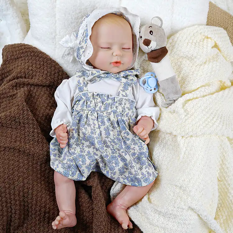 A lifelike reborn doll dressed in a white blouse and blue floral overalls, wearing a matching bonnet, and accompanied by a cute stuffed animal and a pacifier, lying on a cozy blanket
