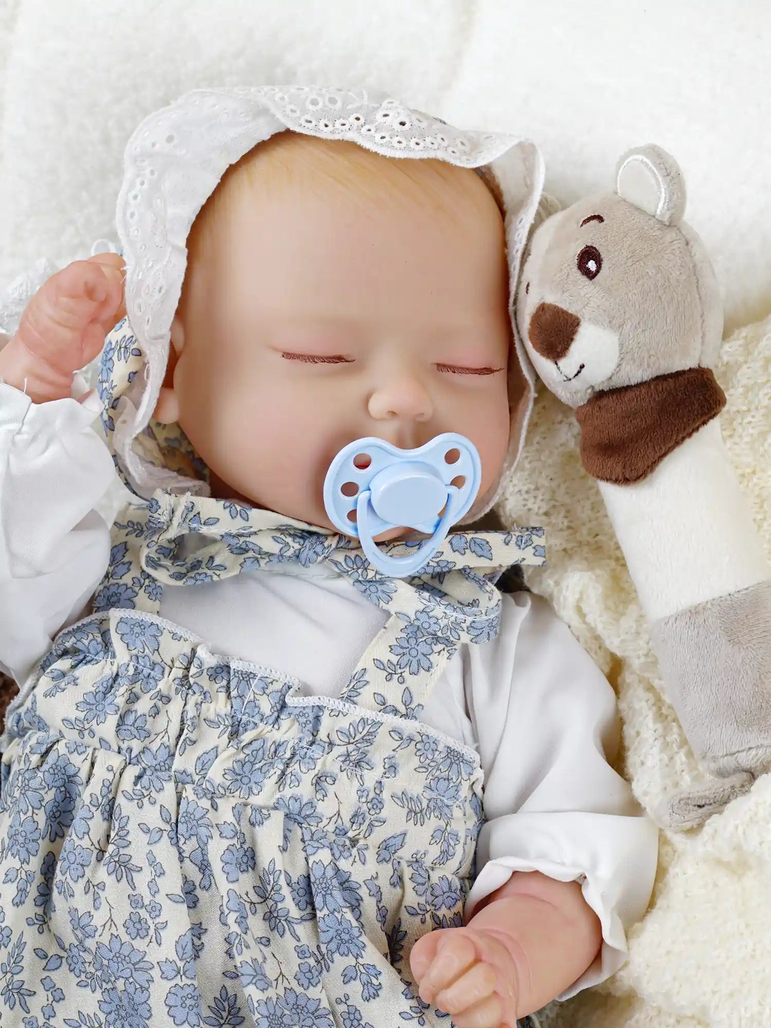 A lifelike reborn doll sleeping peacefully with a blue pacifier in its mouth. The doll is wearing a white and blue floral outfit with a lace bonnet and is holding a plush toy