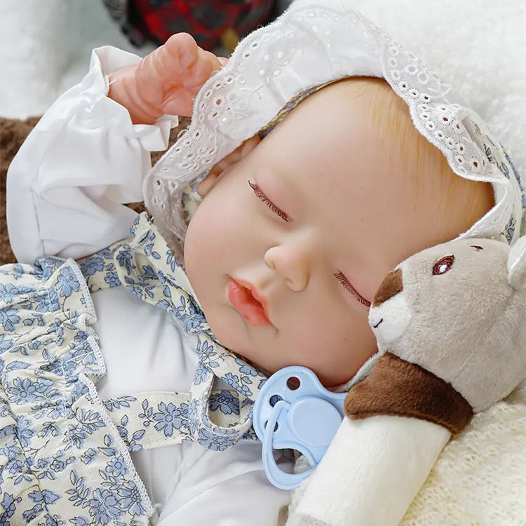 A realistic reborn doll peacefully sleeping, dressed in a white and blue floral outfit with a lace bonnet. The doll is holding a plush toy and has a blue pacifier resting beside it