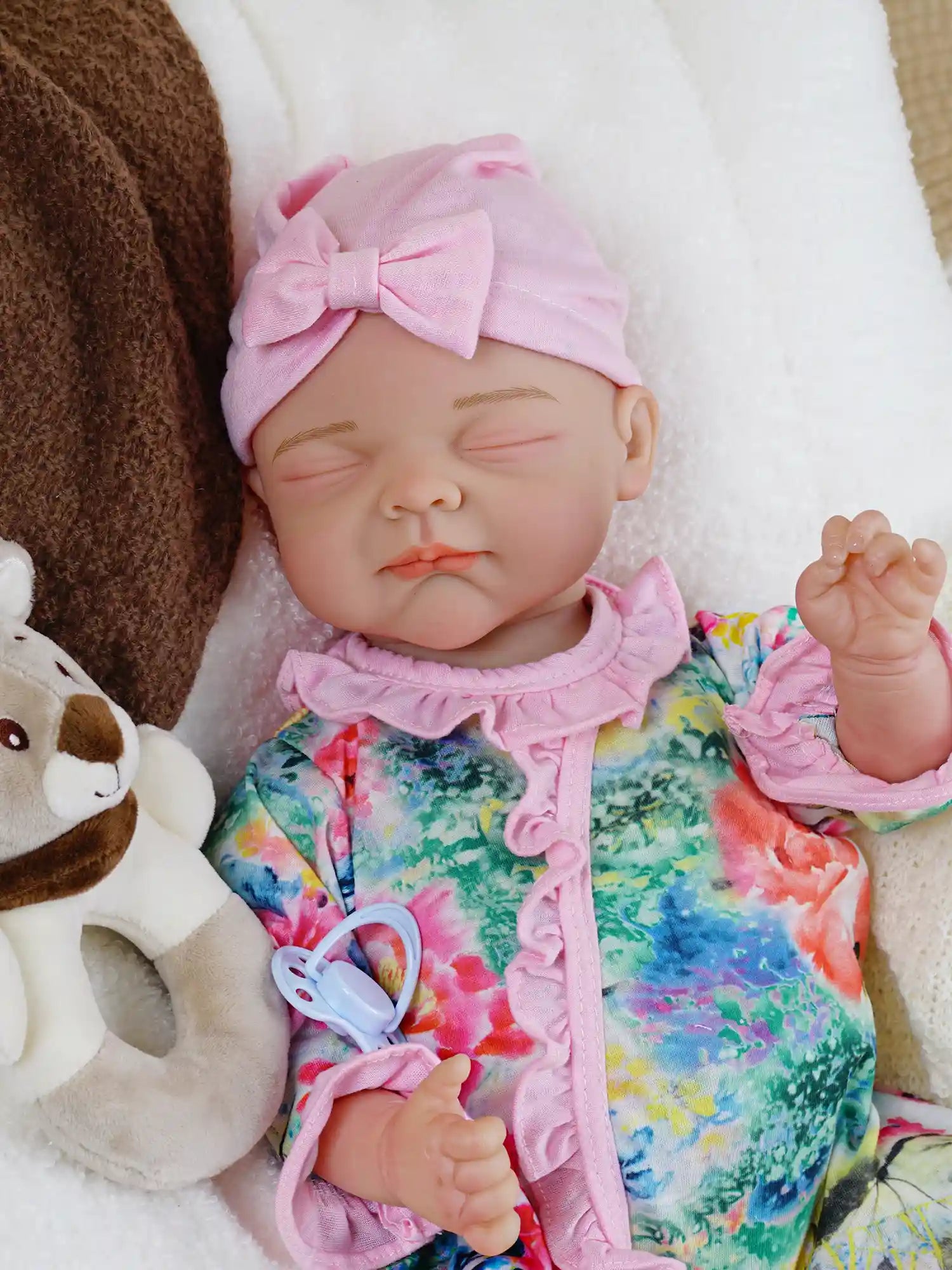A lifelike reborn baby doll wearing a pink bow headband and a colorful floral jumpsuit lies peacefully with closed eyes, holding a white pacifier in one hand.