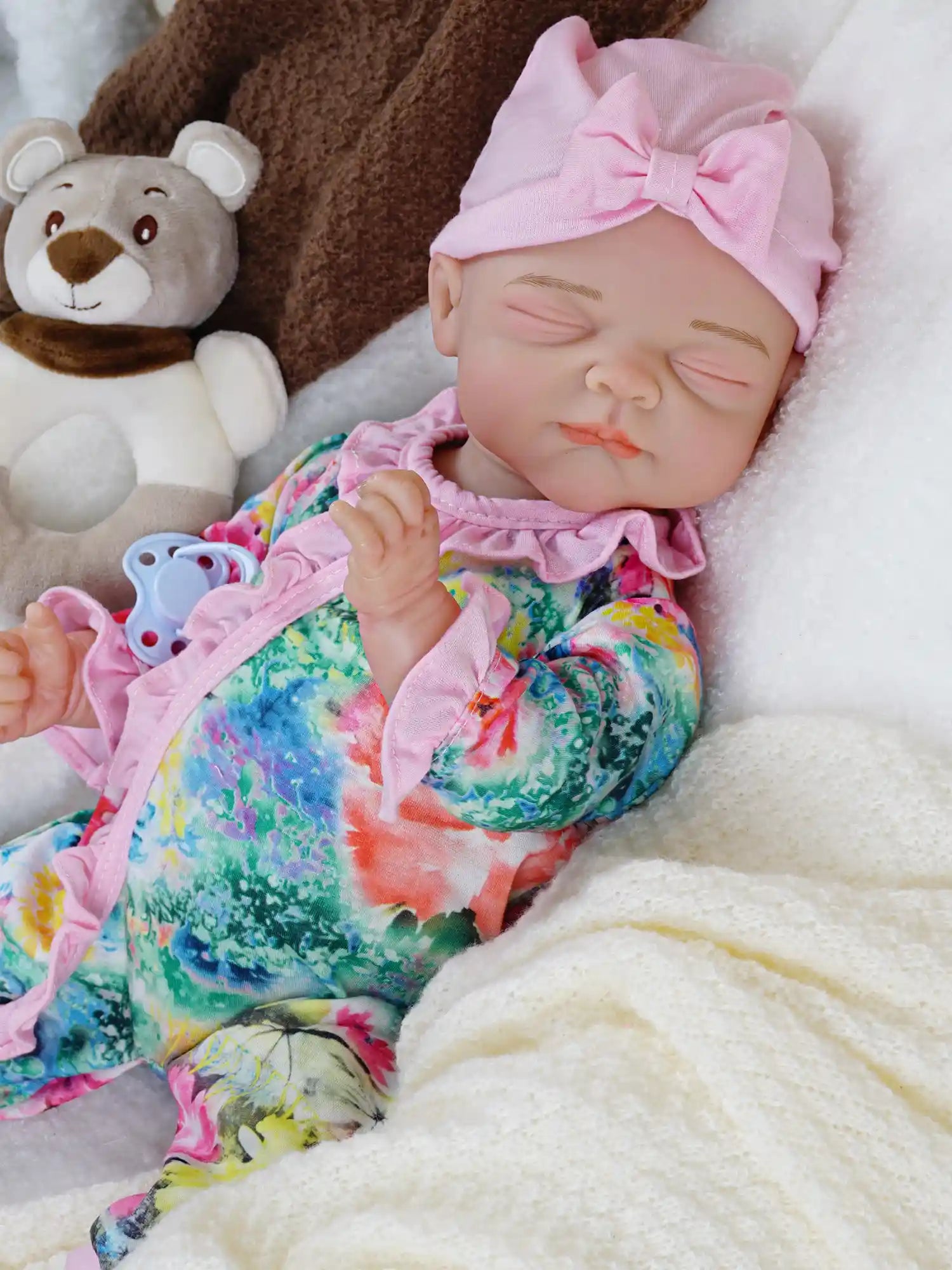 This image shows the same doll as the first, now covered with the cream blanket, the plush teddy bear nearby, and a blue pacifier placed next to her.