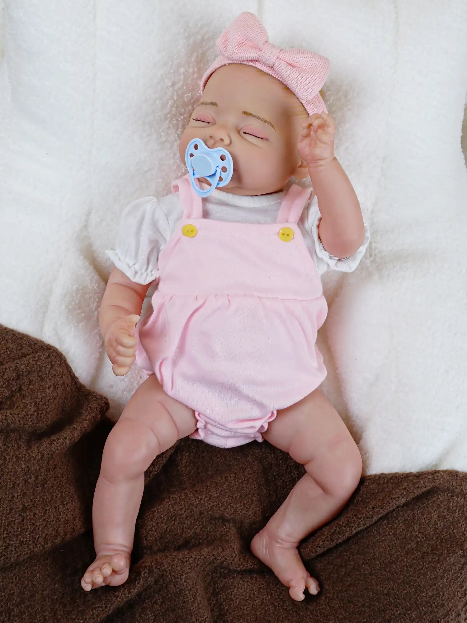 A peaceful reborn baby doll with a soft pink bow headband, gently holding a blue pacifier, lying on a fluffy white blanket.