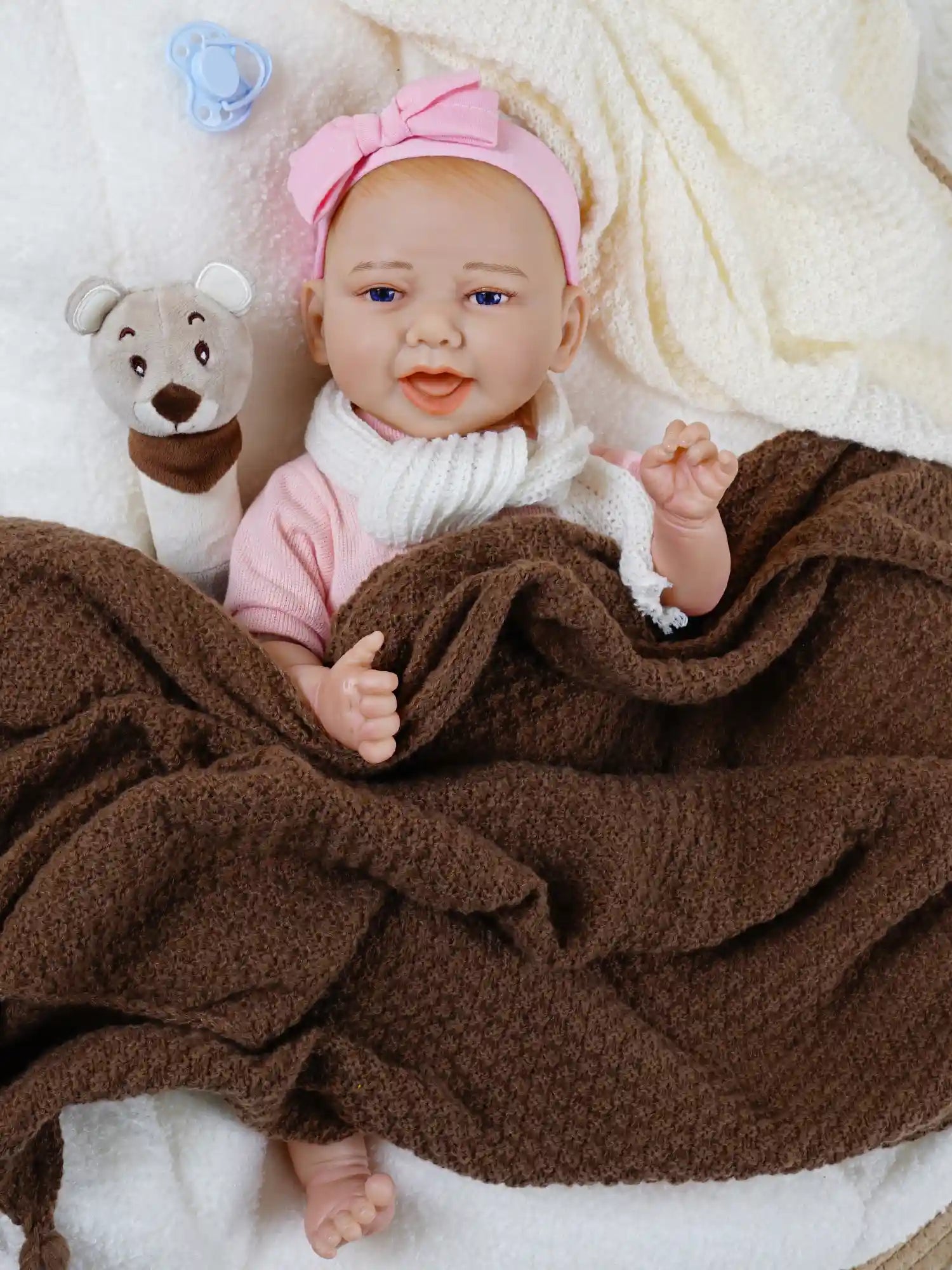 Smiling reborn baby doll wearing a white headband and beige dress, reclining on a white blanket with a brown blanket and plush bear toy, along with a blue pacifier