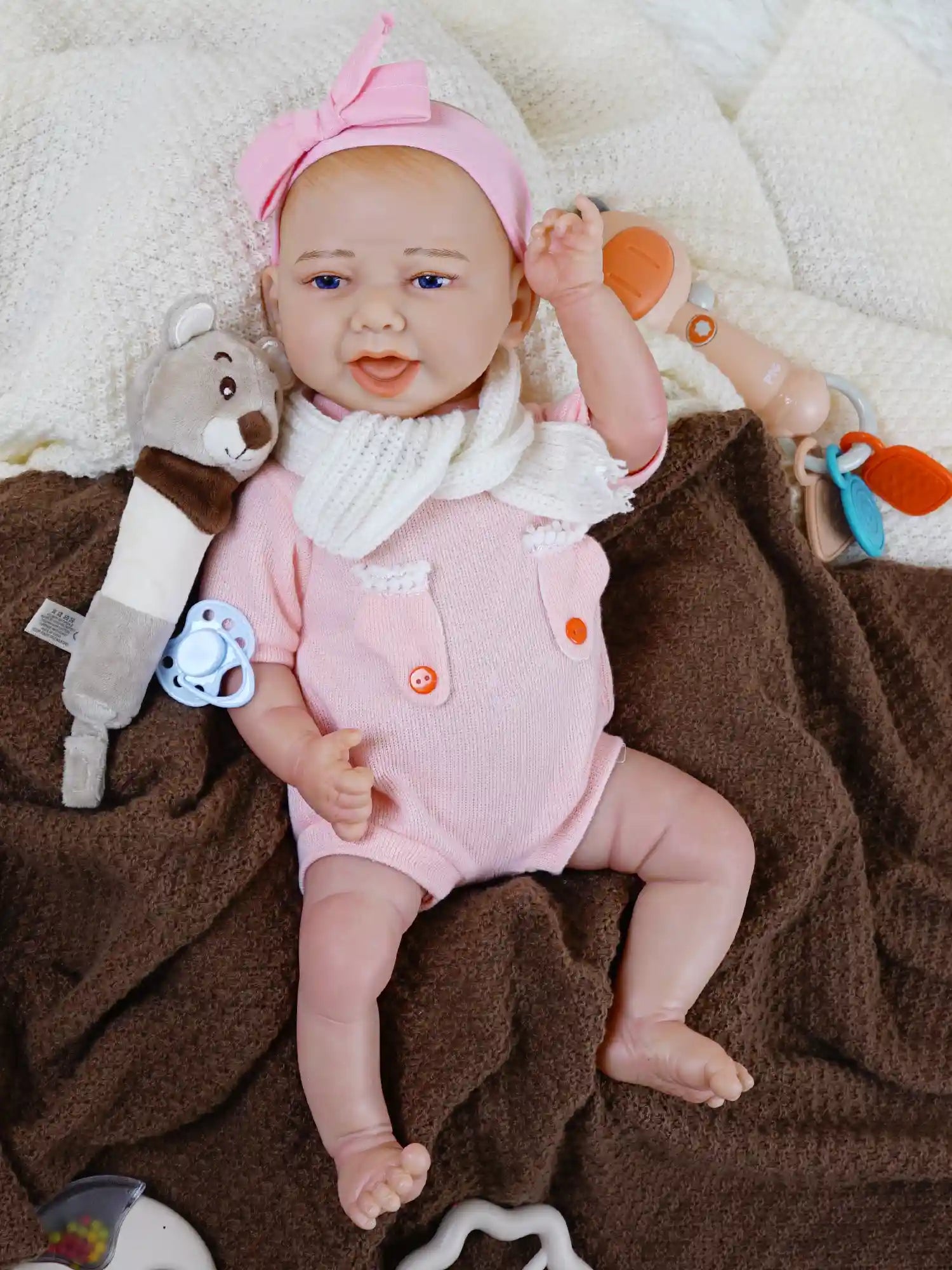 Reborn baby doll with blue eyes and a white bow headband lying on a white blanket, next to a brown blanket, with a plush bear toy and a blue pacifier nearby.