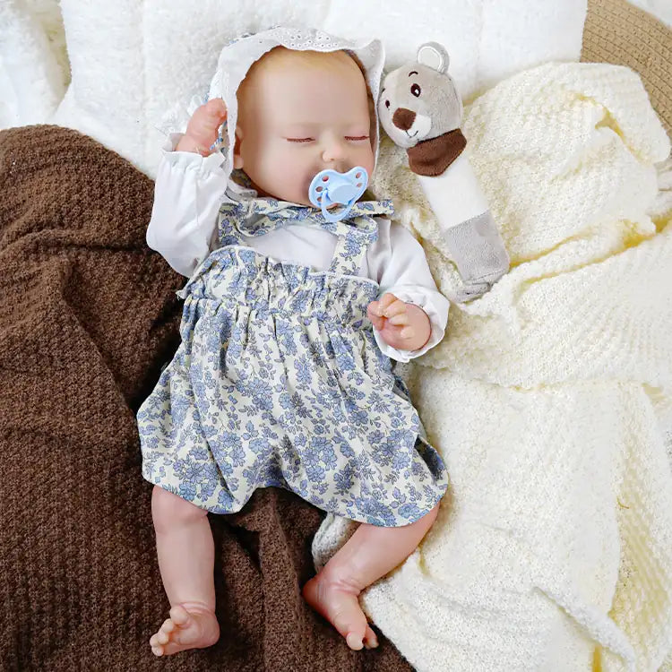 A lifelike reborn doll dressed in a white blouse with blue floral overalls and a lace bonnet, lying on a cozy blanket with a blue pacifier in its mouth.