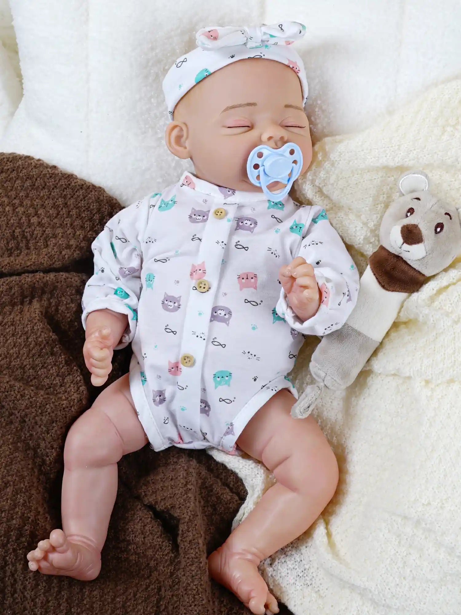 A sleeping reborn baby doll dressed in a white onesie with colorful cat prints, complete with a matching headband and a blue pacifier.