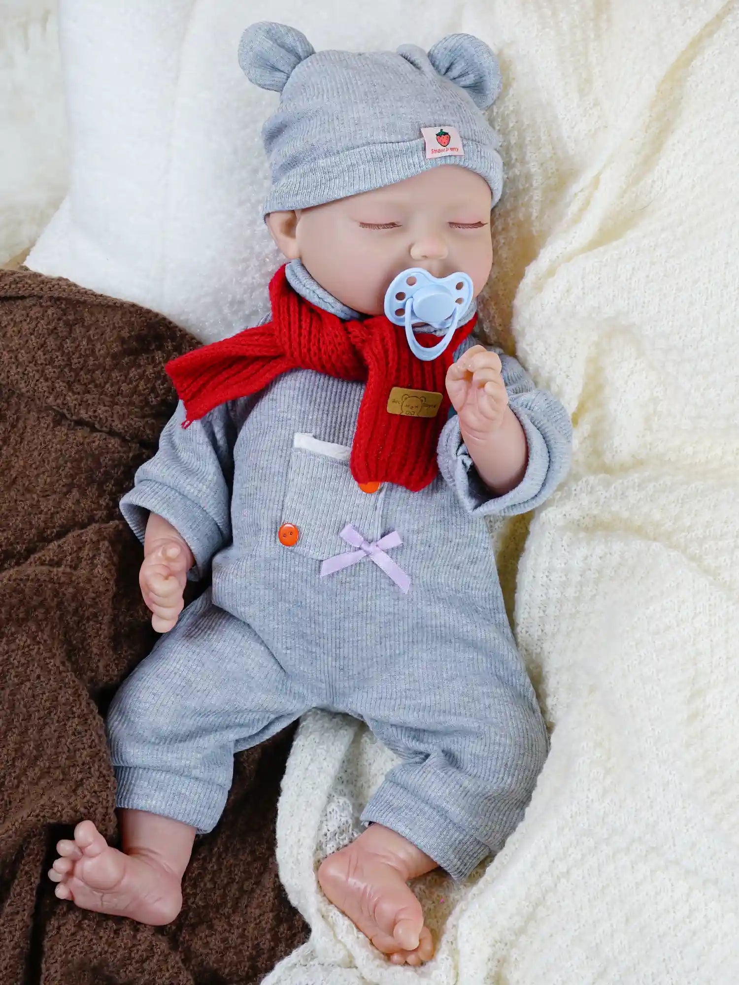 A baby doll dressed in a grey outfit with a matching hat and a red scarf, lying on a soft blanket with a blue pacifier in its mouth and eyes closed, giving it a peaceful, sleeping appearance.