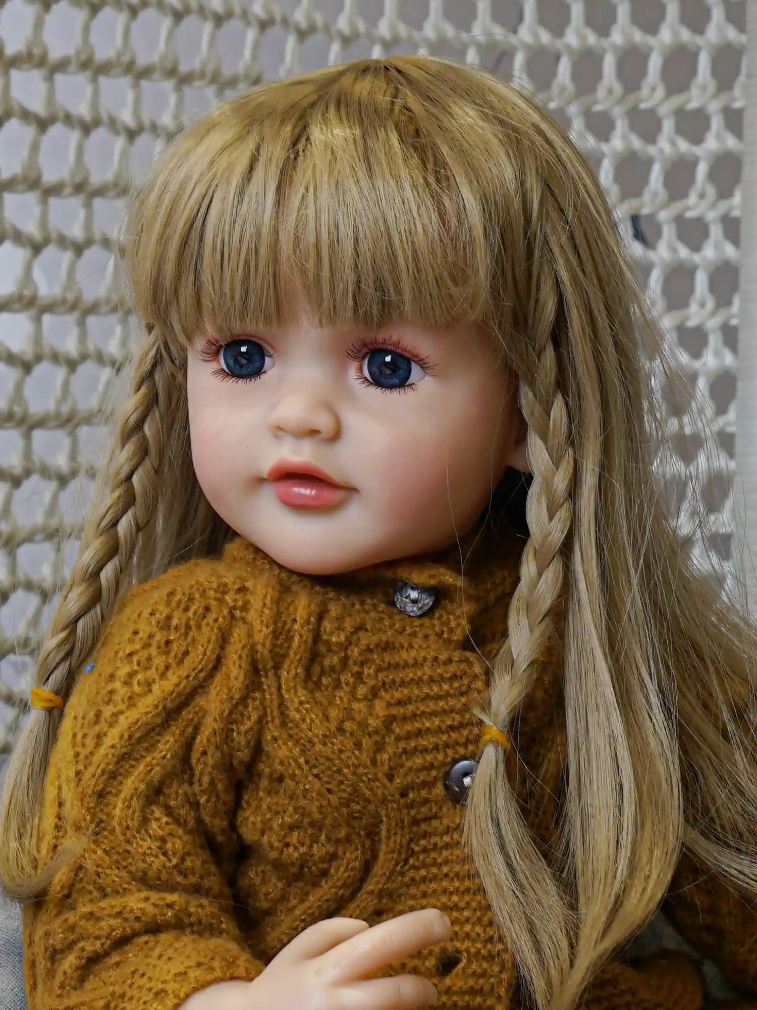 A doll with lifelike features, including blonde braided hair and bright blue eyes, wears a mustard knit sweater and hat, seated against a grey backdrop with a crochet pattern behind.