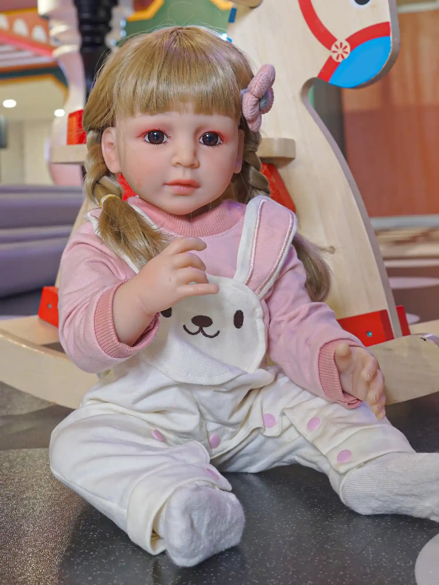 Toddler doll seated on the floor, wearing a pink sweater and white overalls with a bear design, with blonde braided hair and purple hair ties.