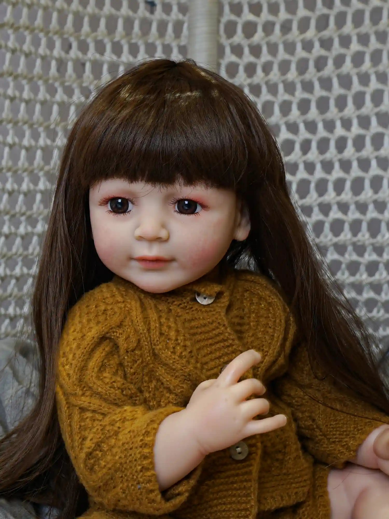 Realistic seated doll with dark brown hair and eyes, wearing a cozy mustard-yellow knit cap and cardigan, with a soft gray cushion as a backdrop.