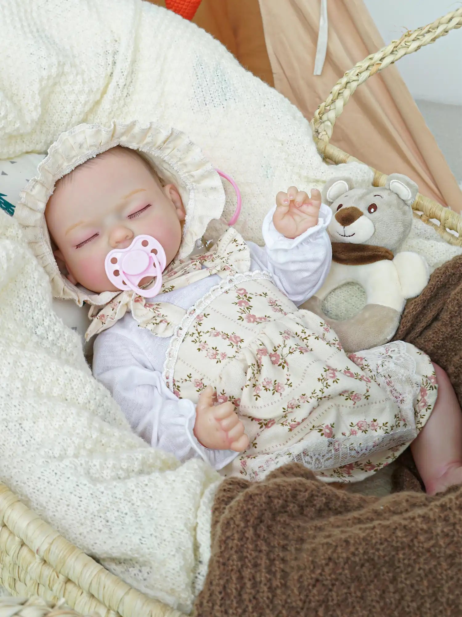 A serene reborn baby doll wearing a floral bonnet and dress, sleeping in a woven basket with a pink pacifier in its mouth, flanked by a soft, knitted blanket and a plush teddy bear.