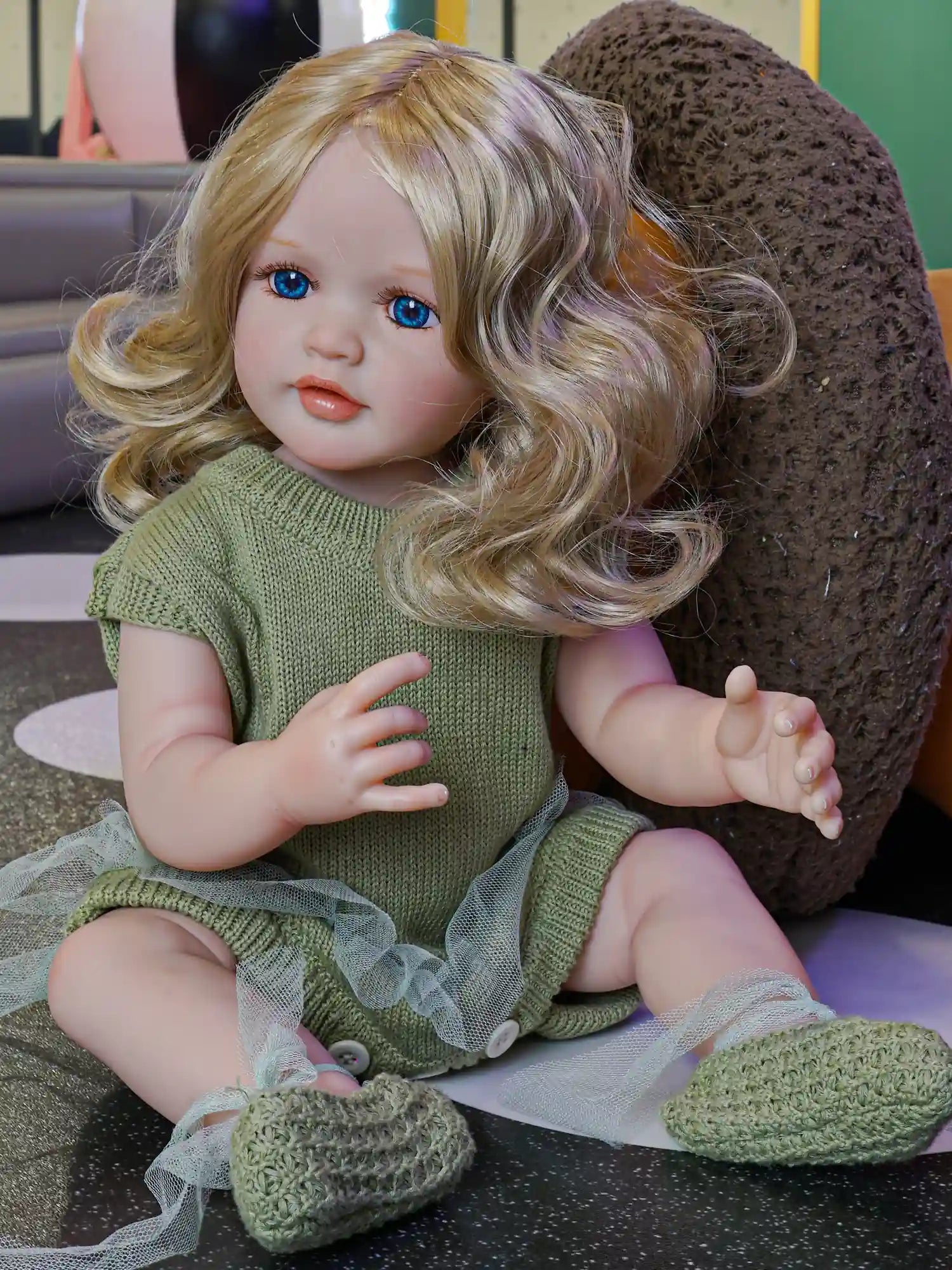 A lifelike reborn doll sitting comfortably, with soft blonde curls and clear blue eyes, dressed in a green outfit with a tulle skirt.