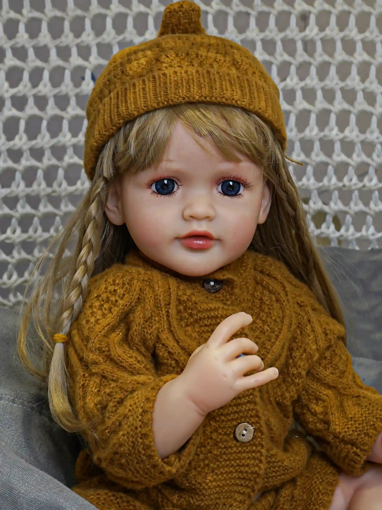 Reborn doll with an intricate facial expression, adorned in a cozy mustard-colored knitted outfit with a matching beanie, set against a textured grey cloth and lace background.