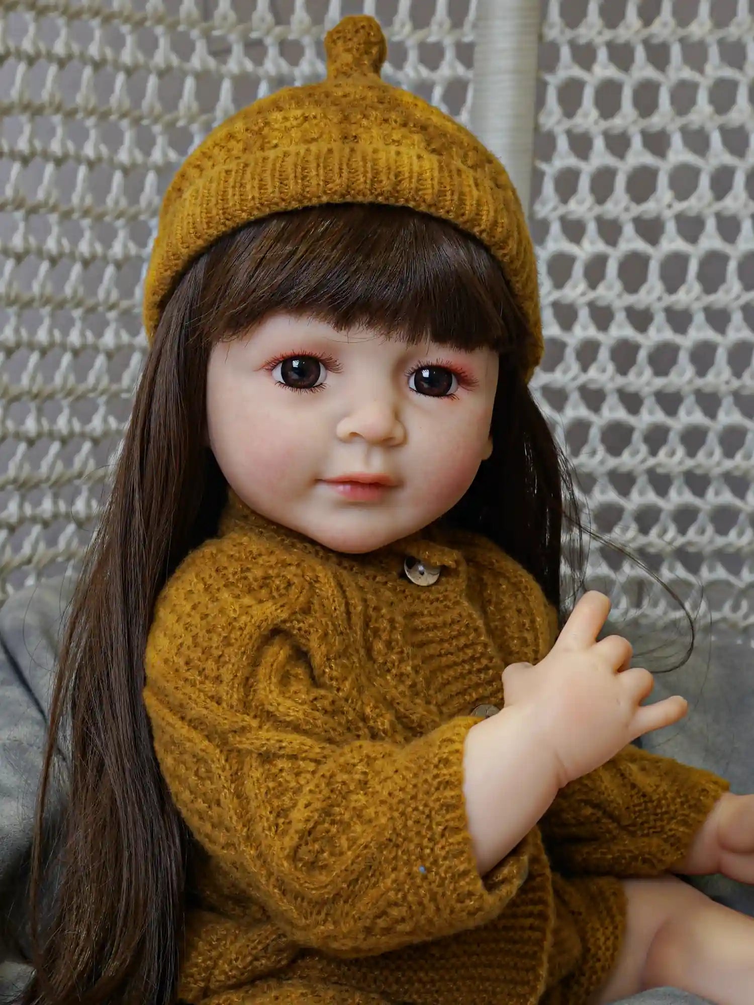 Lifelike doll with long straight brown hair and a knit mustard-yellow beanie, clad in a textured sweater, giving the impression of a contemplative child in autumn.