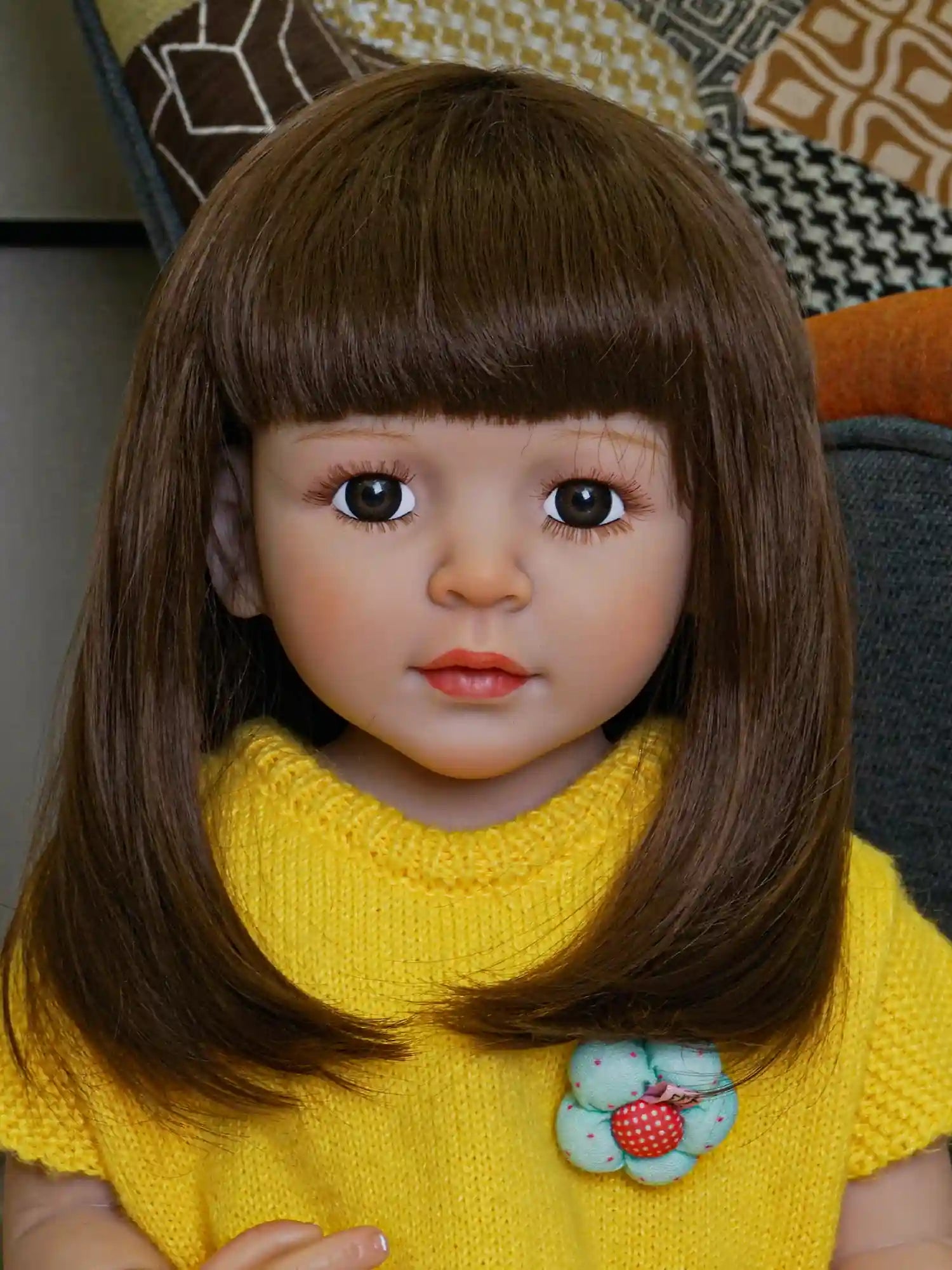Lifelike reborn doll seated on a pink pouf, with smooth brown hair and expressive eyes, wearing a vibrant yellow knit dress with a playful tulle hem.