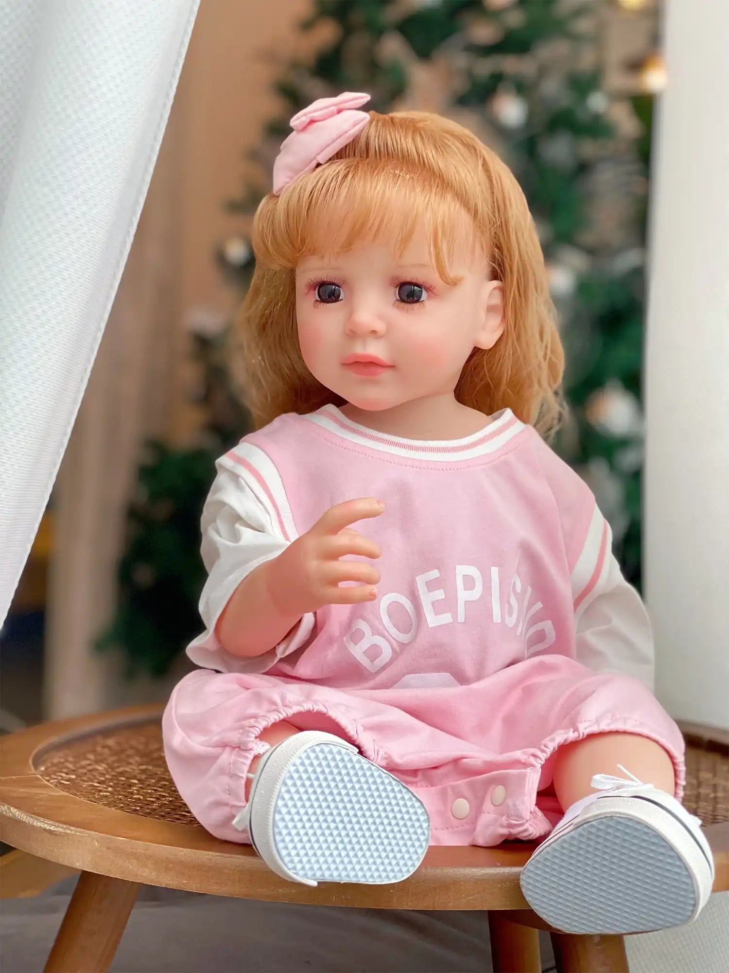 Lifelike doll seated on a woven chair, sporting a casual pink outfit, with a festive Christmas tree in the background.