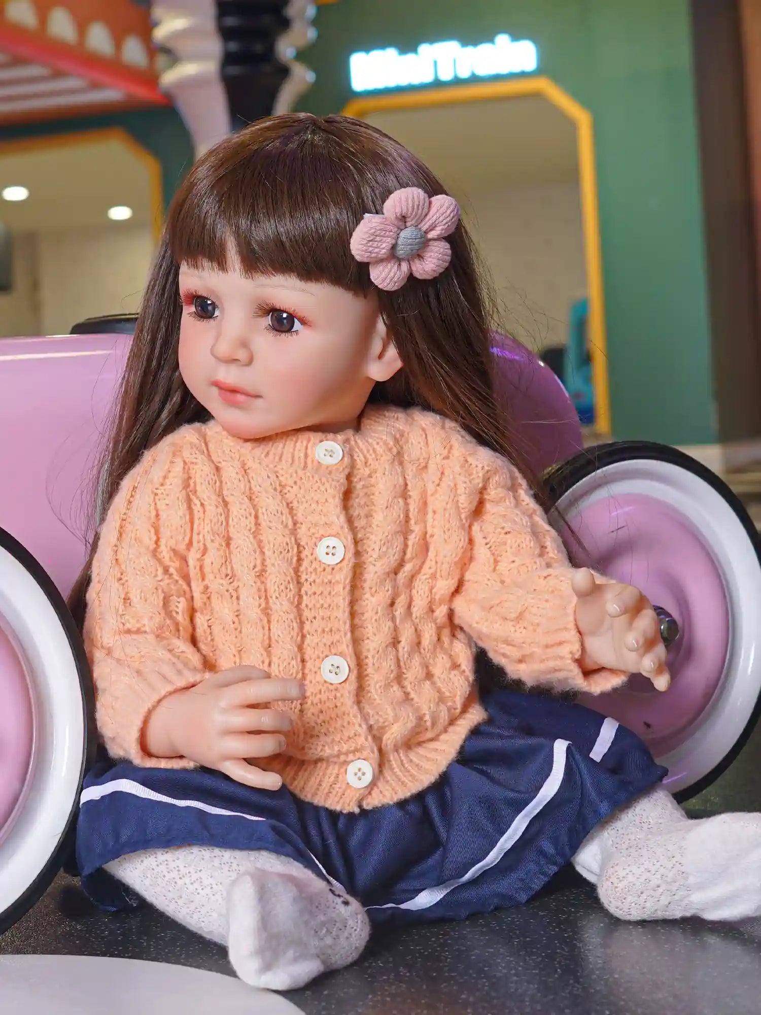 Toddler doll with brown hair and a knitted pink flower hair accessory, wearing an apricot sweater, seated beside a pink toy car.