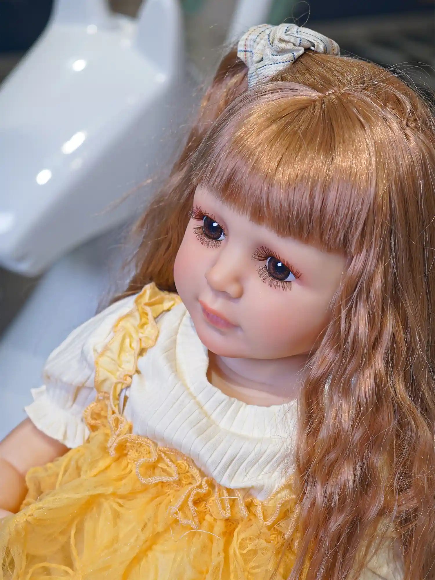 A realistic doll in a vibrant yellow dress, seated on the floor, exuding the innocence of youth with a charming toy car backdrop.
