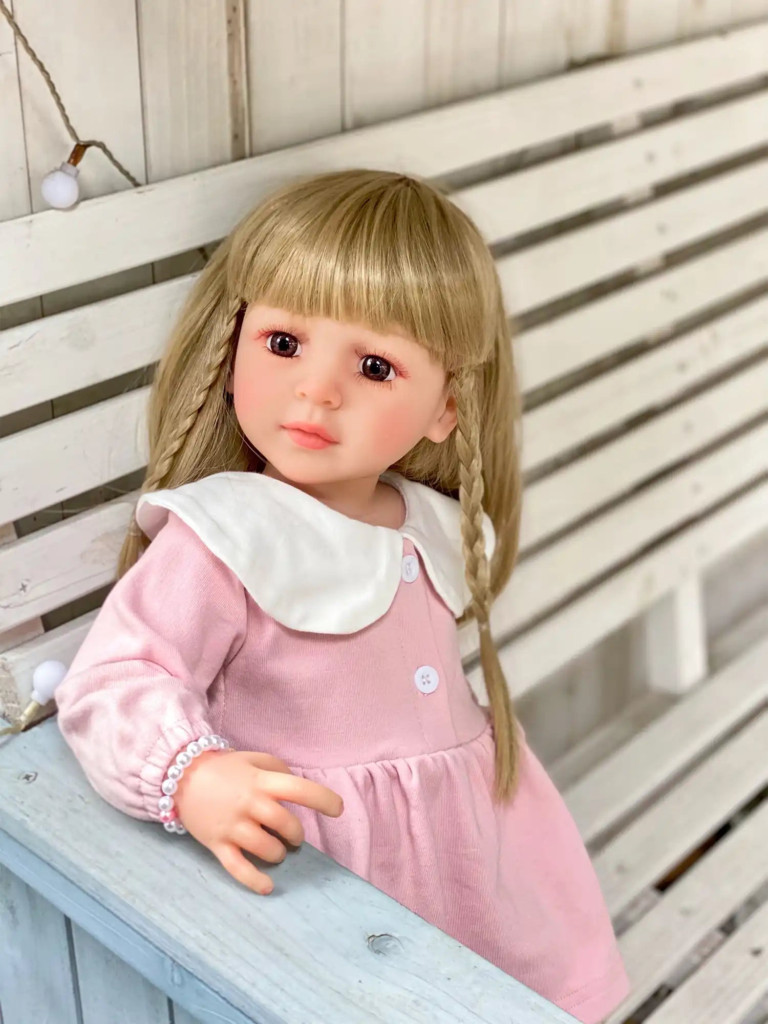 Reborn doll with blonde braids and a pink dress, seated on a rustic bench.
