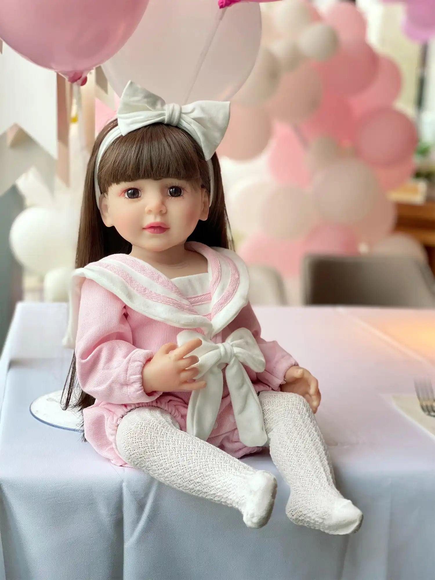 Lifelike doll with dark hair and a white bow, sitting on a wooden bench.