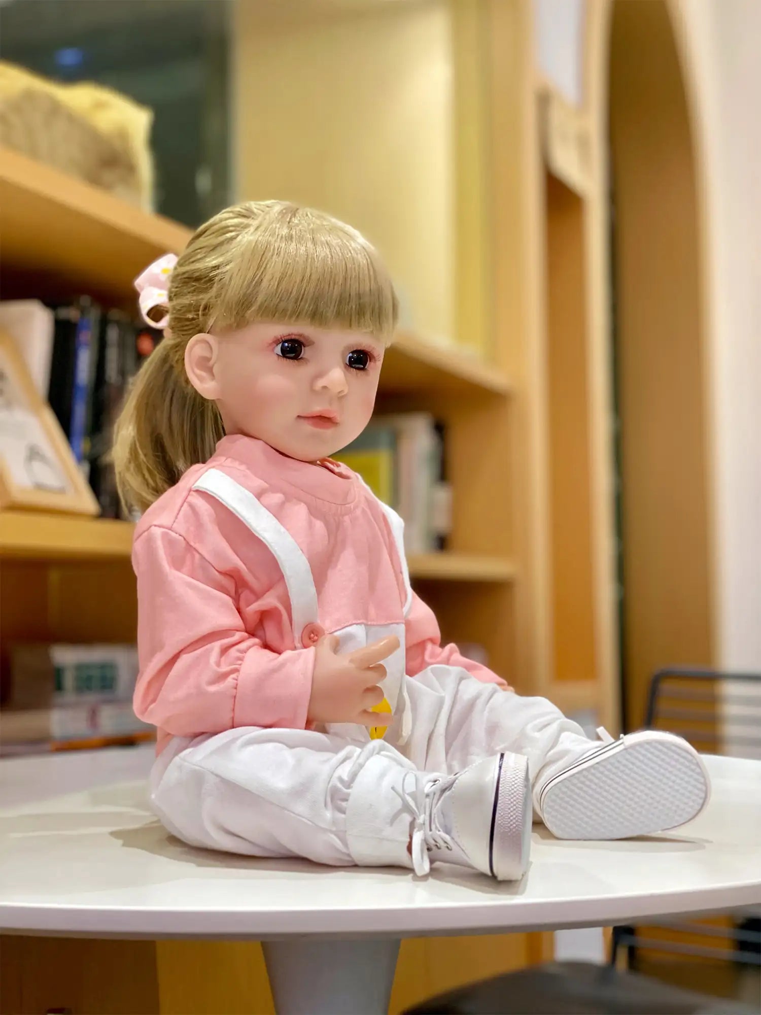 Toddler doll with blonde hair, seated on the floor in a pink sweatshirt and white pants, holding a yellow object, with a toy car in the background.