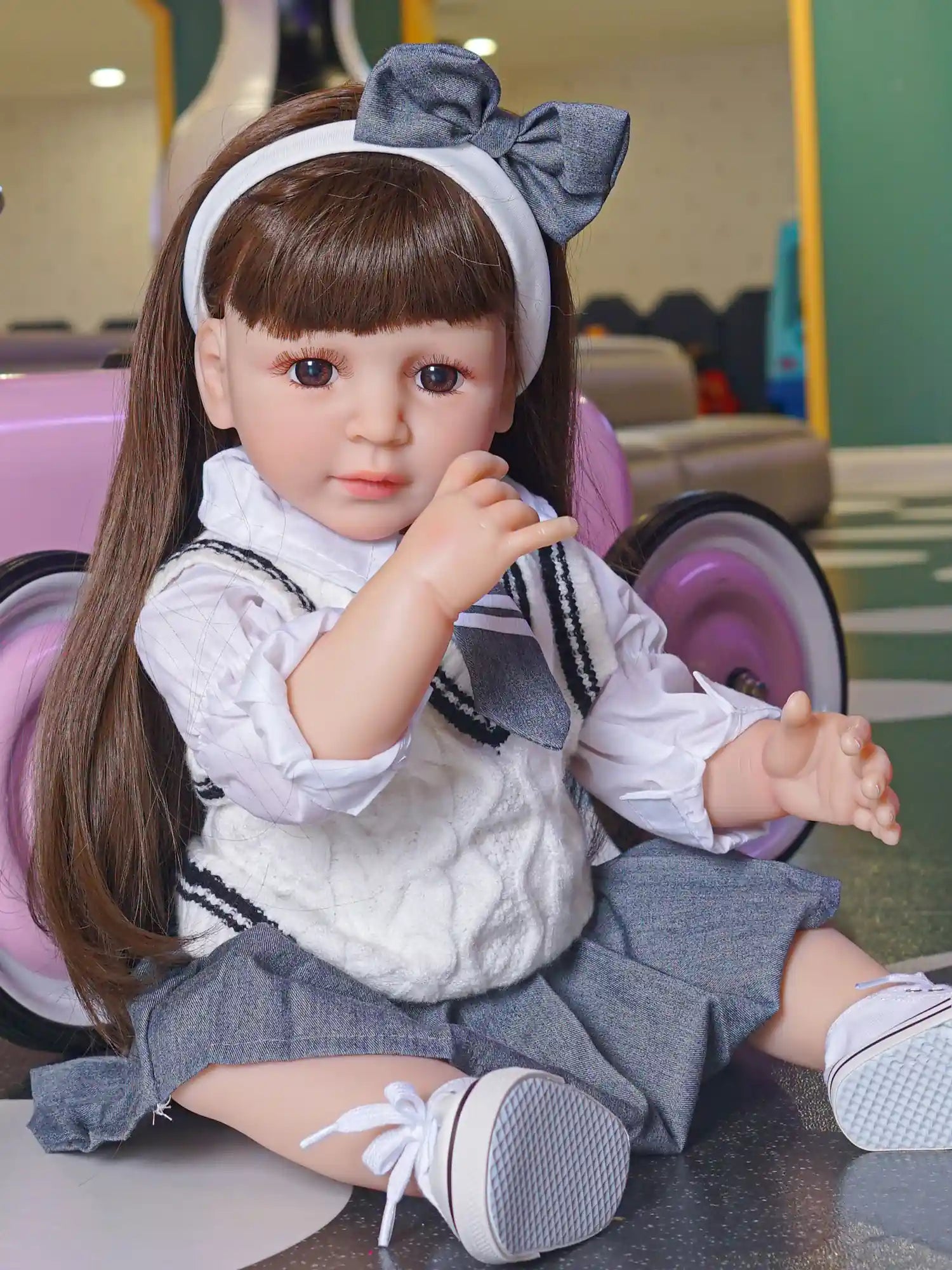 A hyper-realistic doll with long, straight brown hair and brown eyes, sitting with one hand up, wearing a grey and white school uniform, complete with a grey bow headband.
