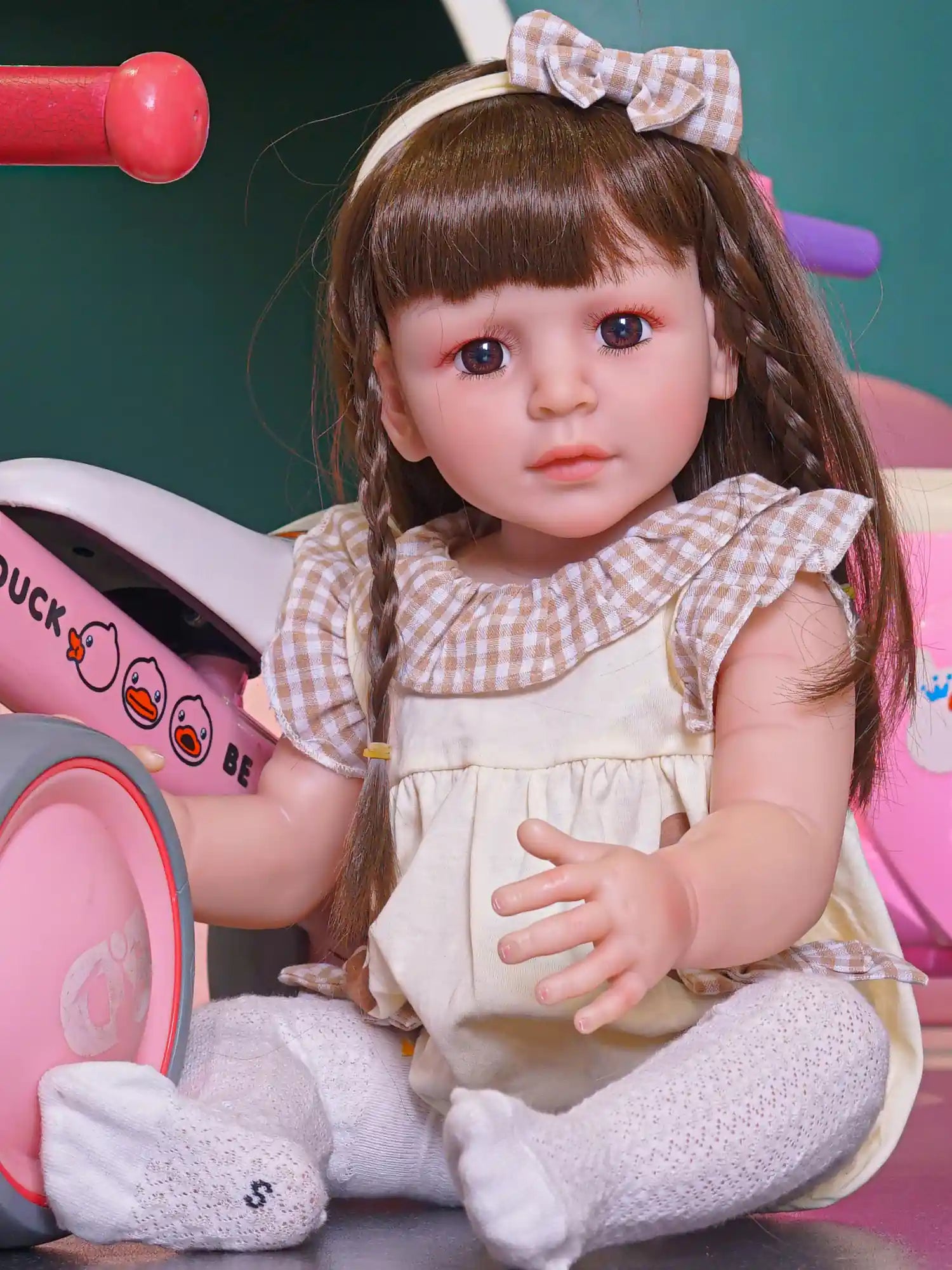 Toddler doll with brown hair and plaid bow, seated with a toy scooter, wearing a cream dress and white tights.