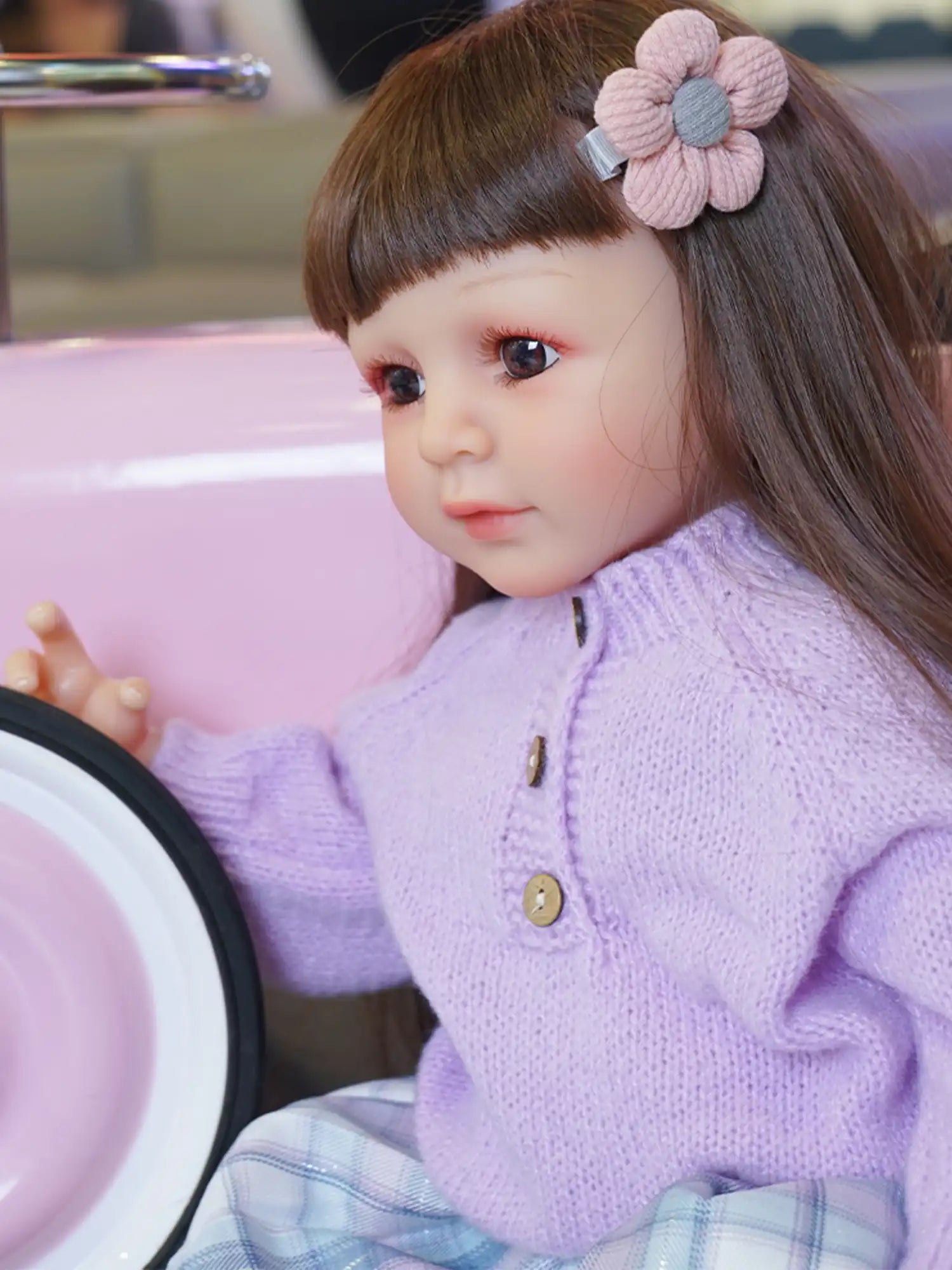 Realistic child doll with sparkling eyes, wearing a cozy purple sweater and checkered skirt, seated indoors.